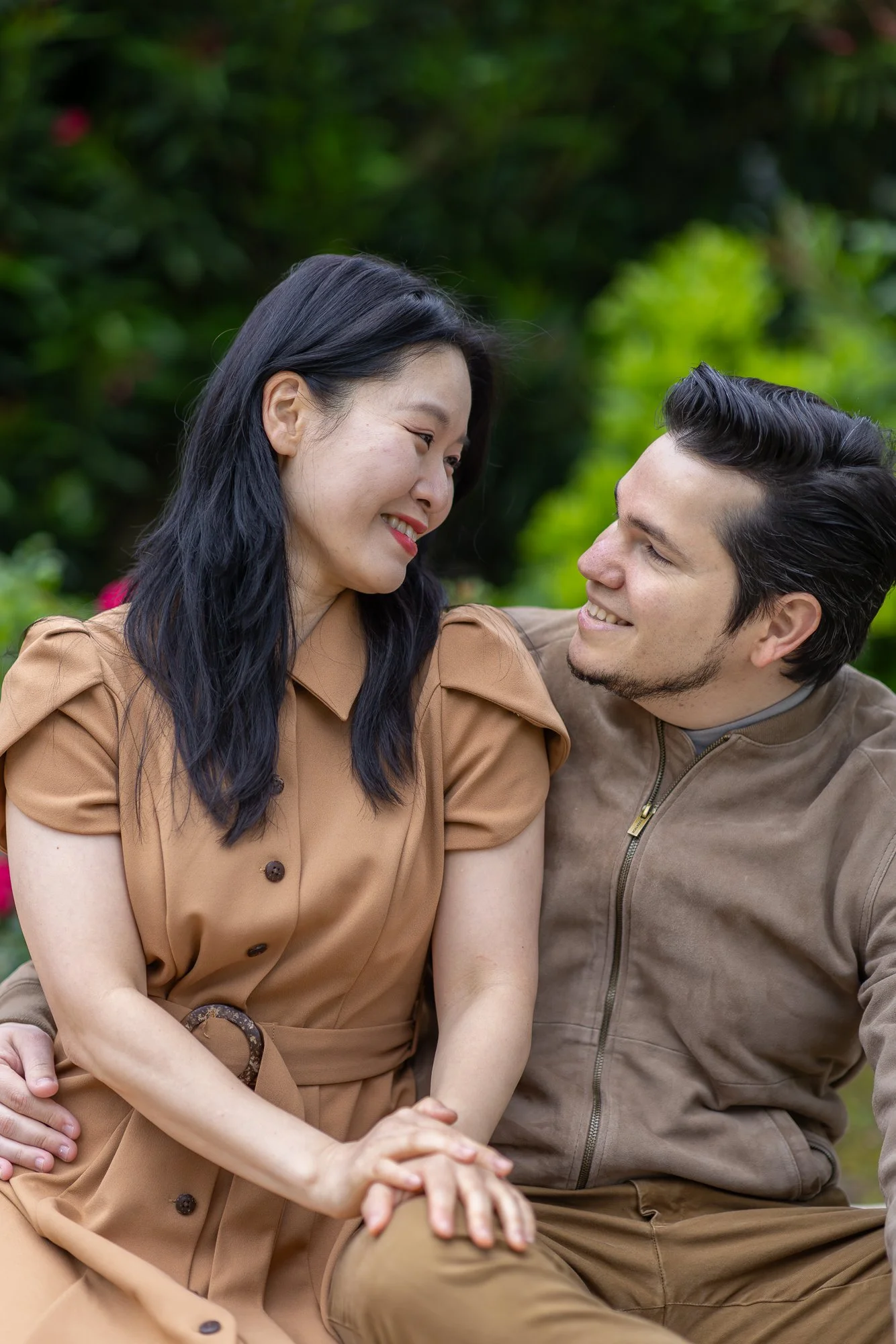 A woman and a man sitting close together outdoors, smiling and looking into each other's eyes, surrounded by green foliage.
