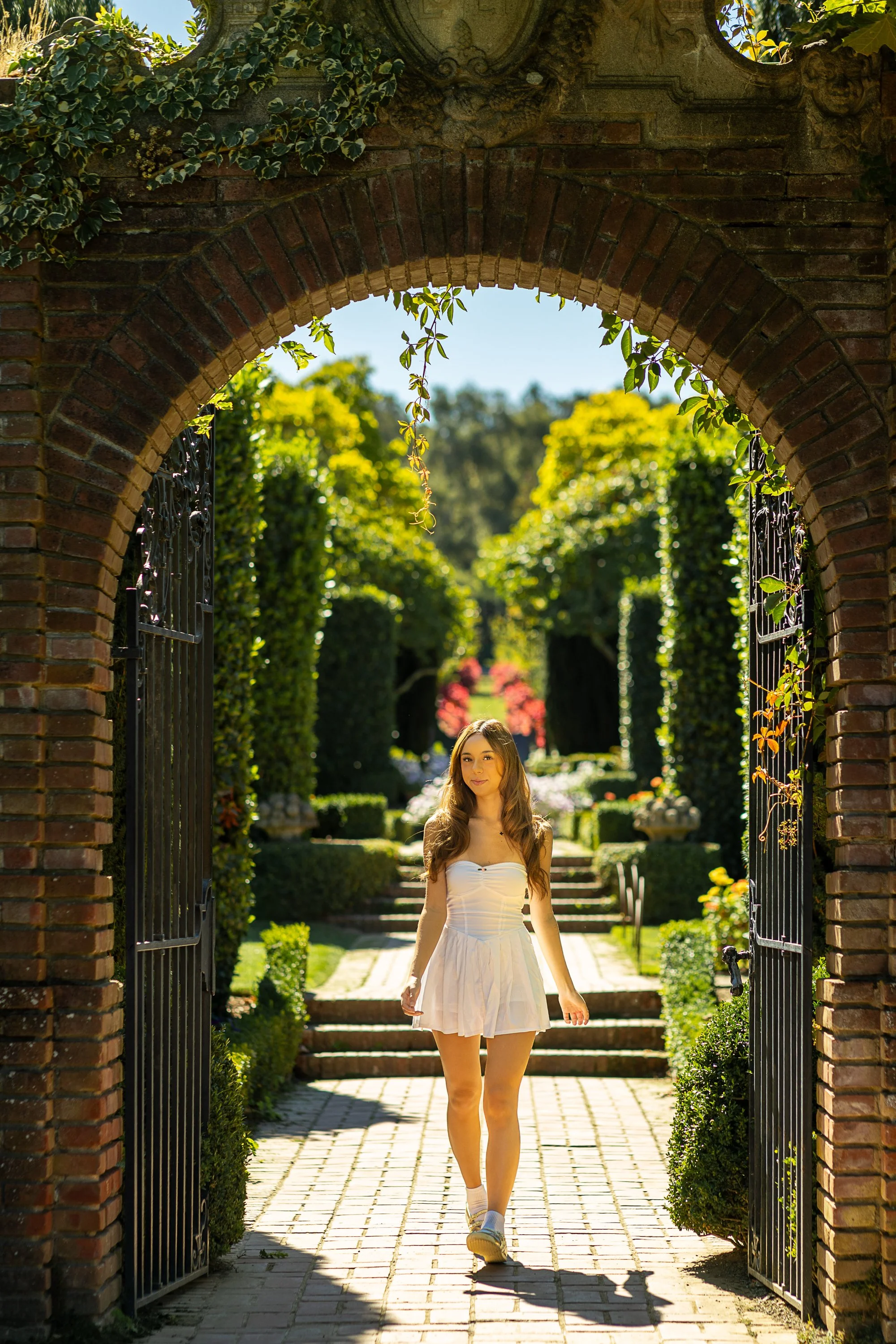 Graduate in white dress framed by brick arch and iron gate at Filoli garden entrance