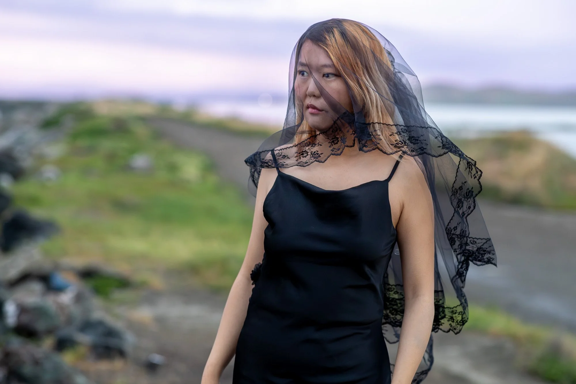 Woman in black dress holding black lace veil open wide with direct gaze, purple pink dusk sky, Dumbarton Bridge Bay Area lifestyle photography