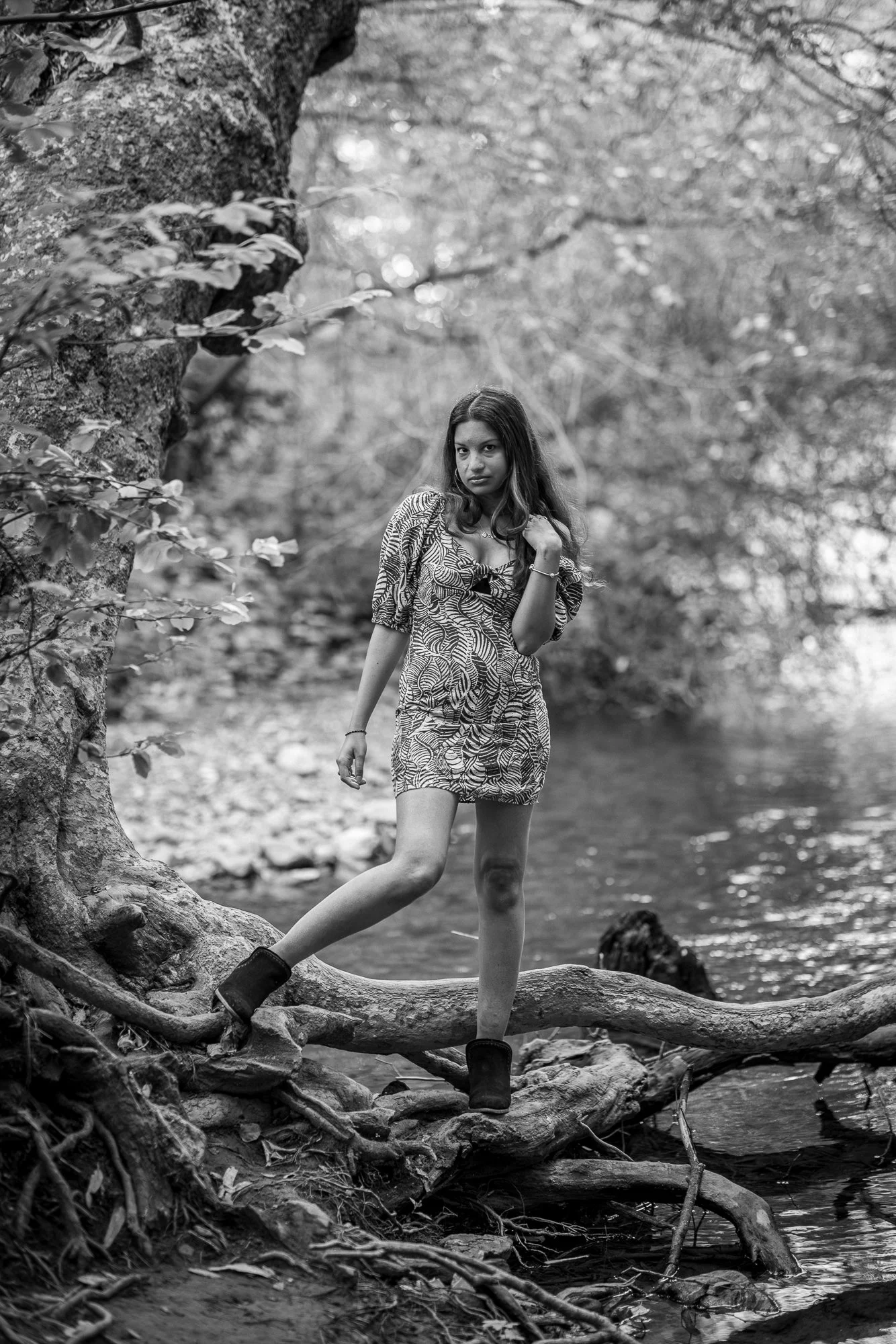 A woman standing on a large fallen tree by a river in a forested area, with trees and leaves in the background.