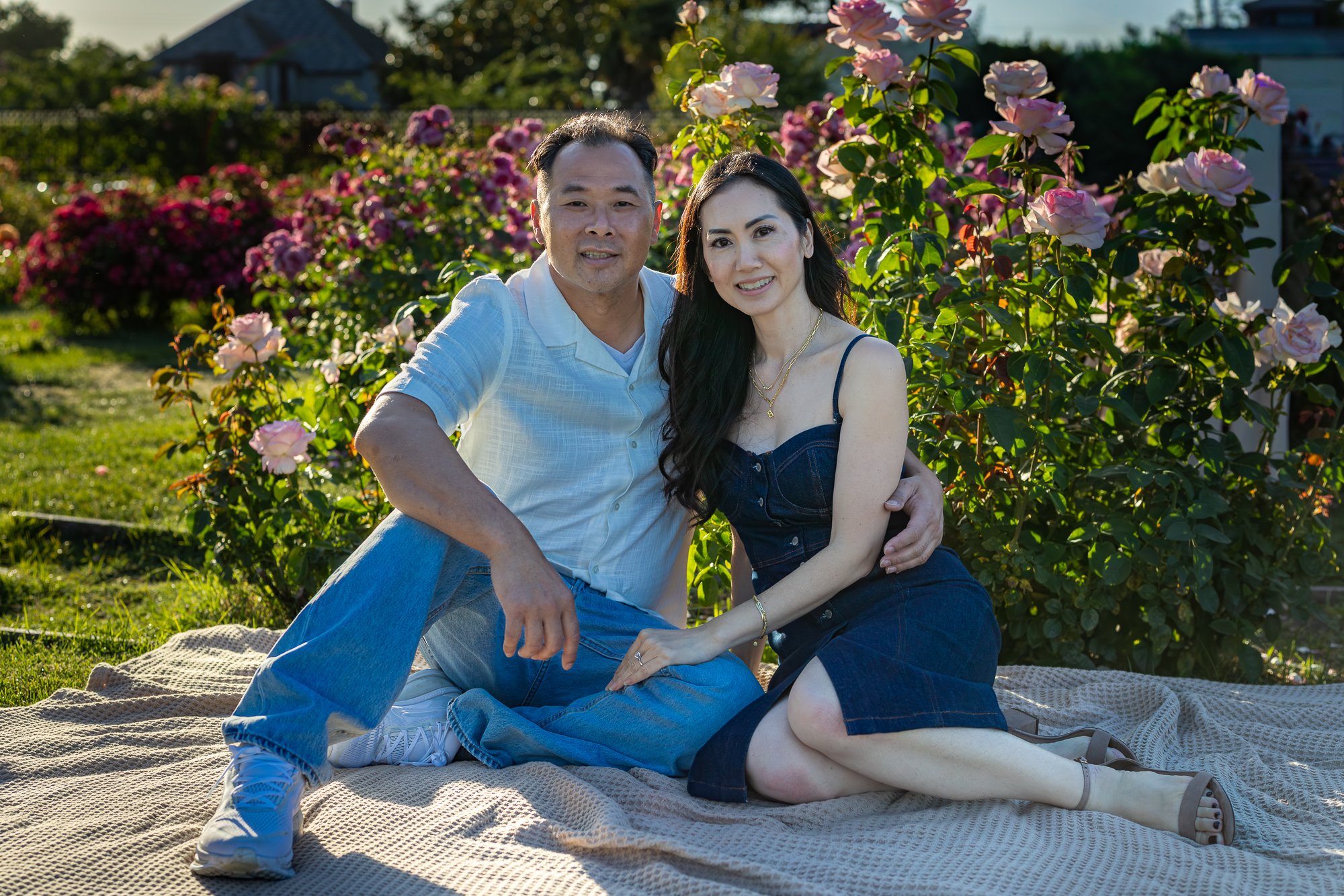 A couple sitting on a blanket in a garden with pink and purple roses, smiling at the camera during sunset.