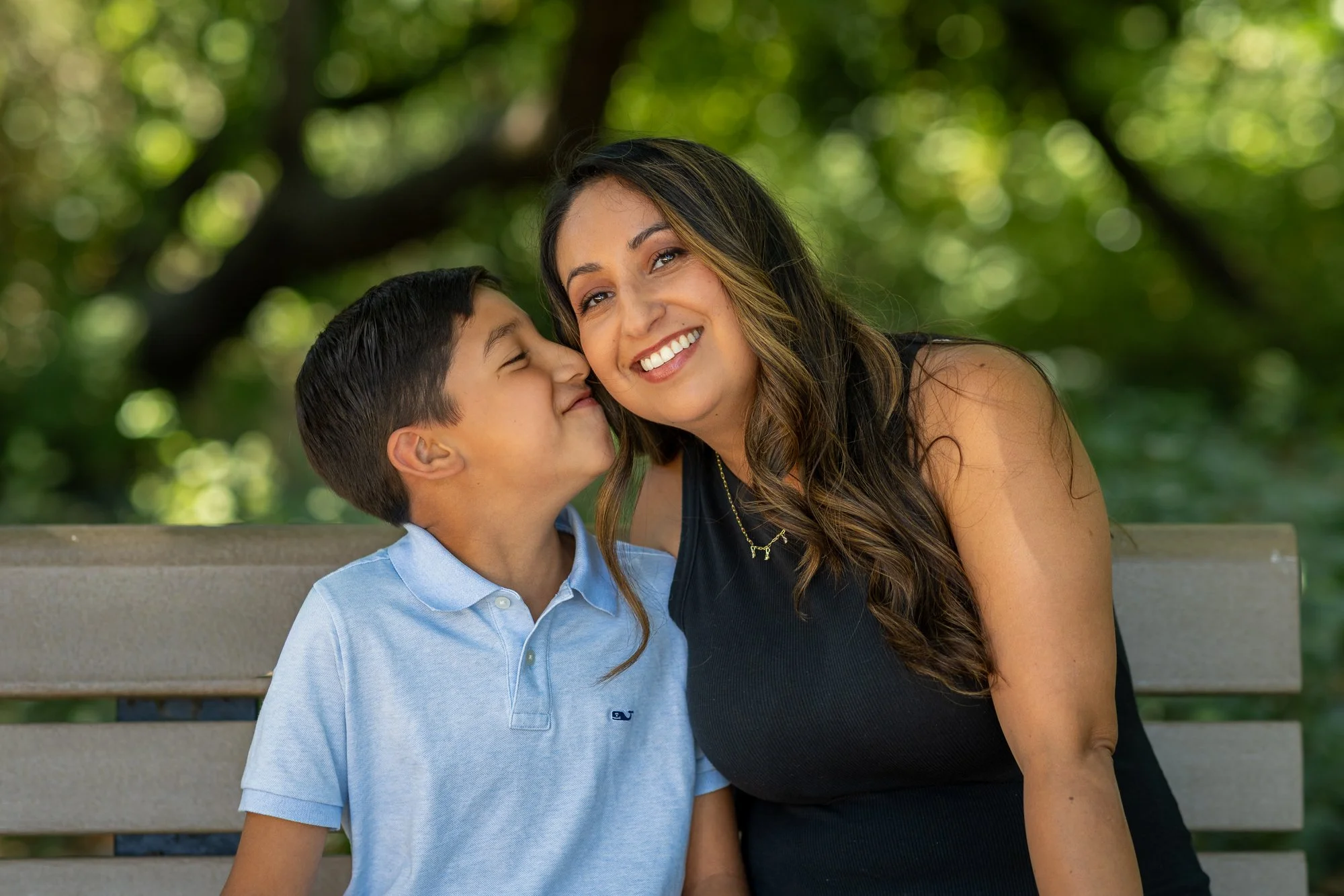 A woman smiling with a boy whispering into her ear on a park bench.