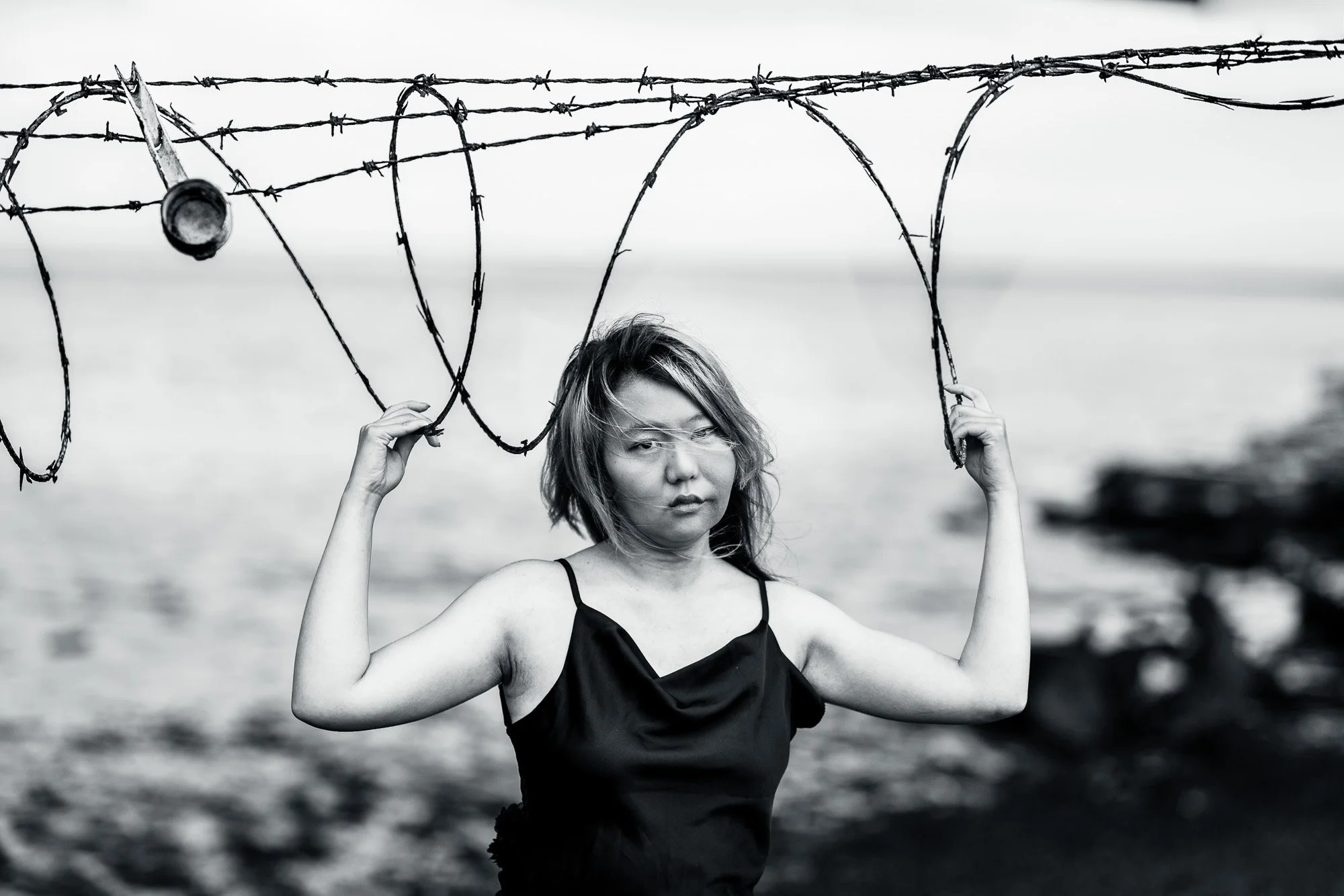 Black and white portrait of woman holding razor wire with direct intense gaze, San Francisco Bay behind, Dumbarton Bridge Shoreline Trail photography