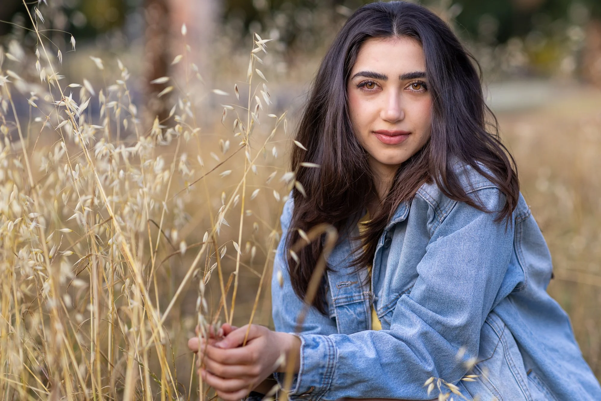 A young woman with long dark hair and brown eyes, wearing a denim jacket, sitting outdoors in a field of tall, dried grasses.