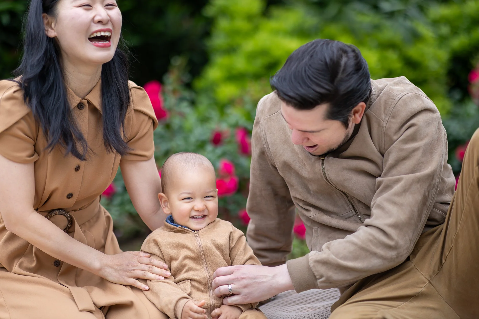 A joyful family moment with a woman, a man, and a smiling toddler in an outdoor garden with pink flowers.