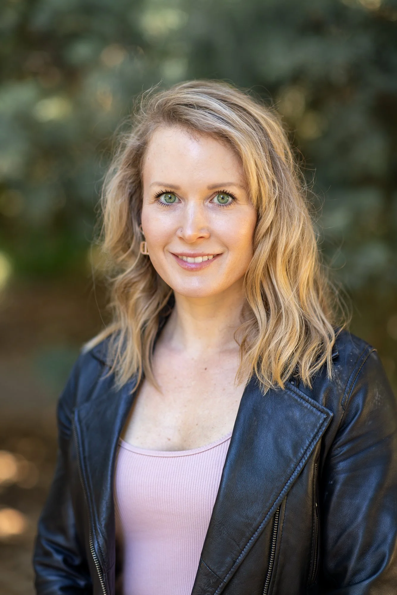 A woman with wavy blonde hair and green eyes smiling outdoors, wearing a black leather jacket and a light pink top, with blurred green trees in the background.