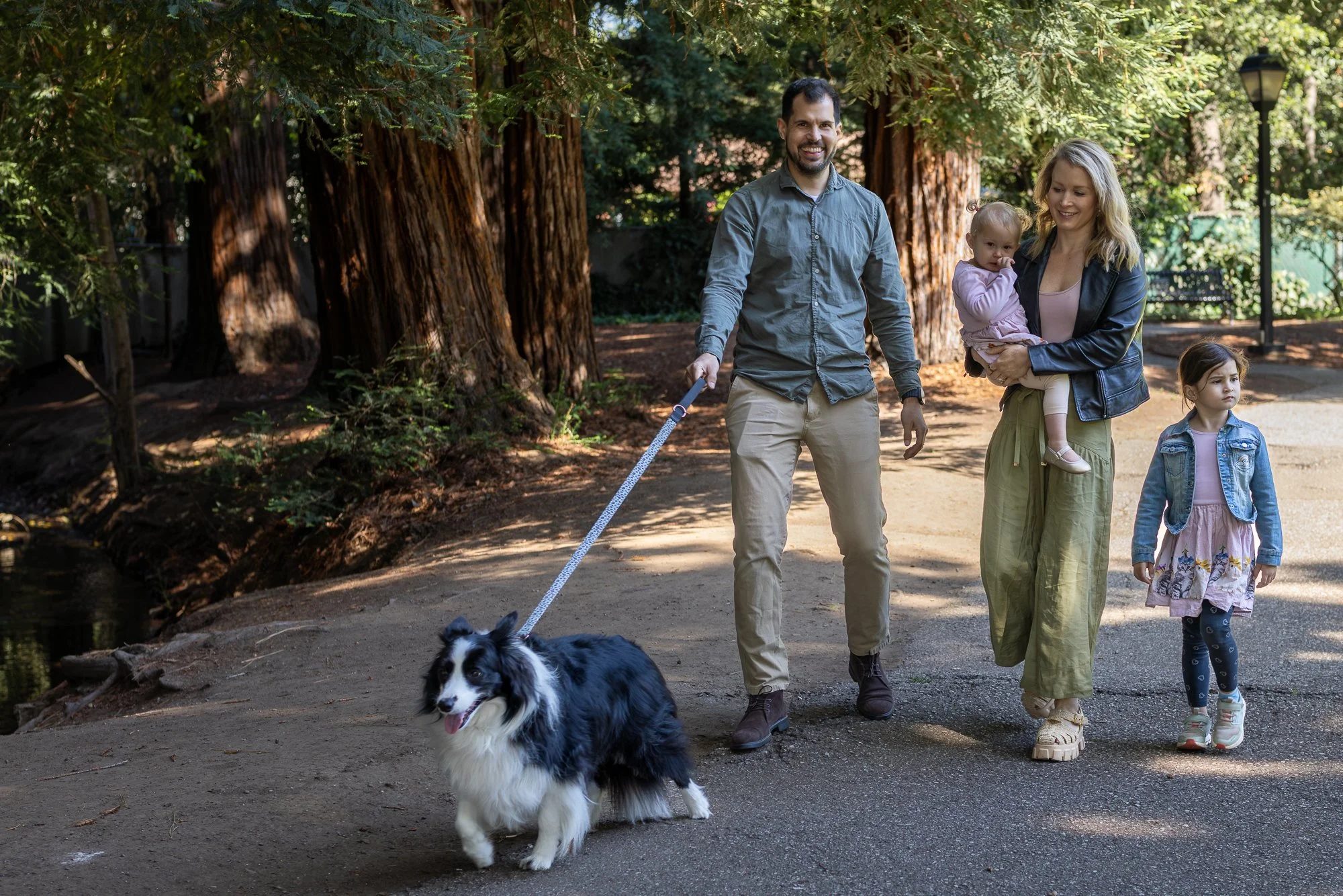A family of four walking their dog in a park surrounded by tall trees. The father is holding the dog's leash, and the mother is carrying a young girl who is biting her finger. Another girl is walking beside them. The park has greenery, benches, and a