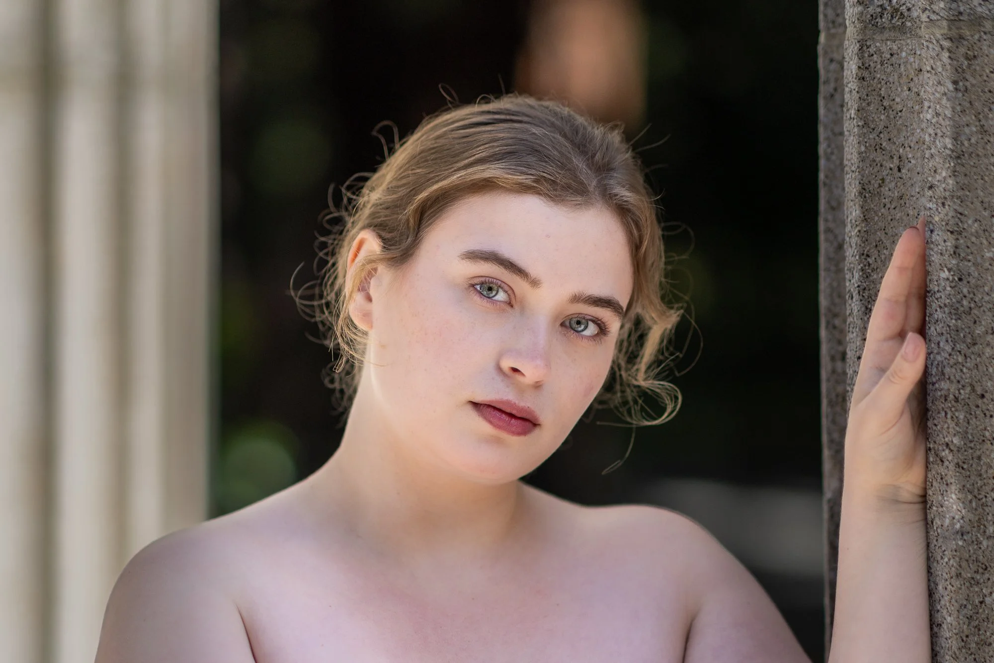 A young woman with light hair and blue eyes looks directly at the camera while standing outdoors near a concrete wall, touching the wall with her right hand.