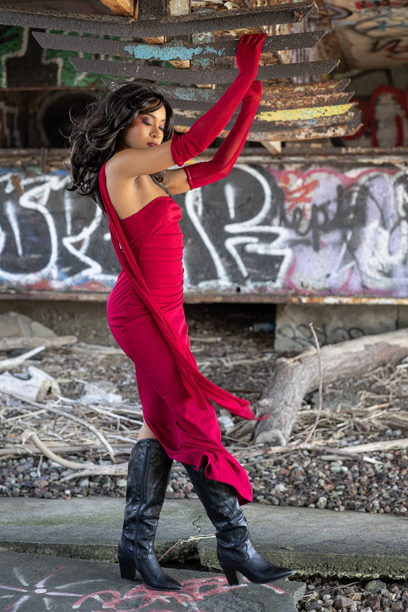 A woman with dark hair wearing a red dress with long red gloves and black high-heeled leather riding boots, standing on a sidewalk in front of a graffiti-covered wall and graffiti on the pavement.