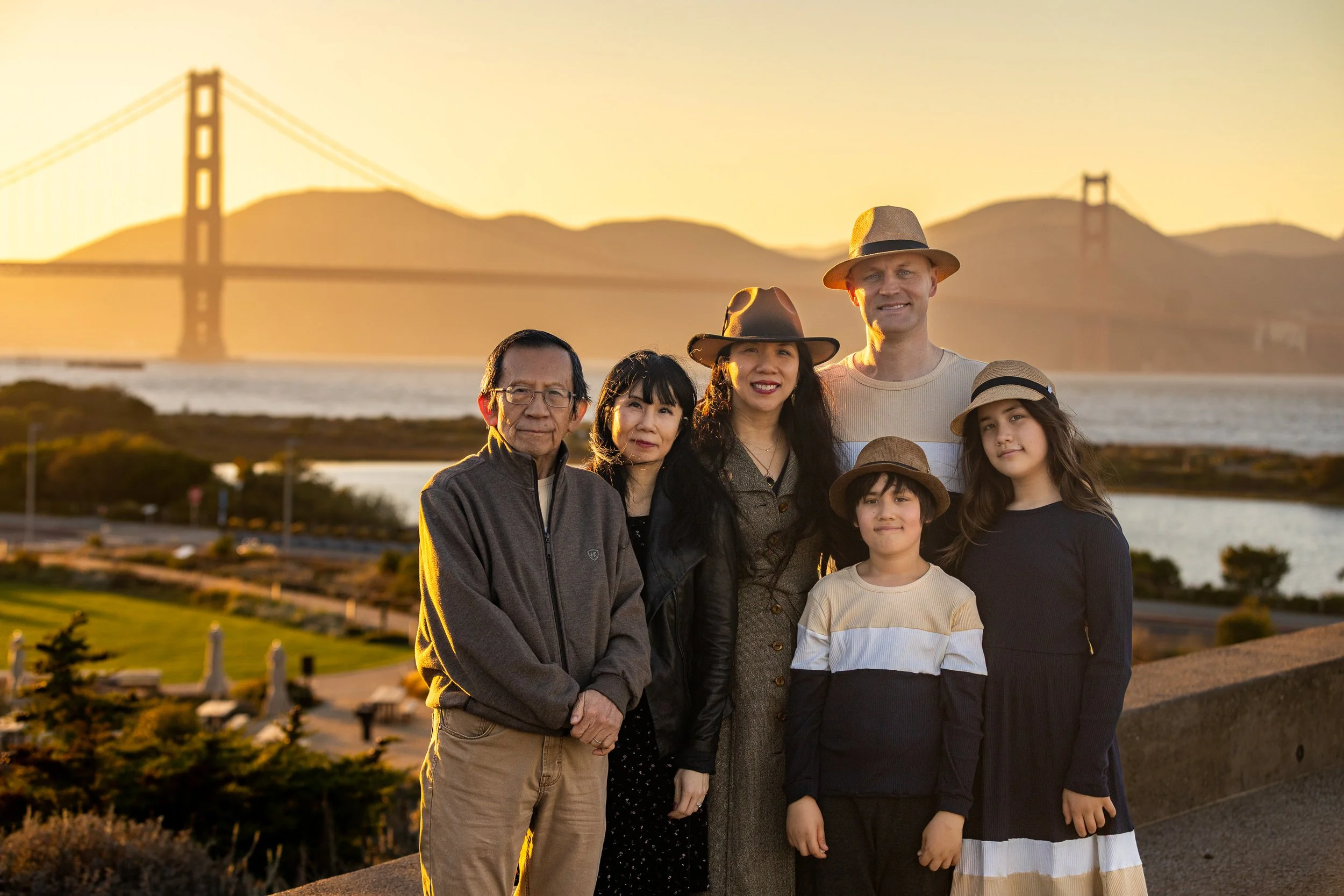 A diverse family of seven people standing outdoors at sunset with the Golden Gate Bridge in the background, smiling and wearing casual clothing and hats.