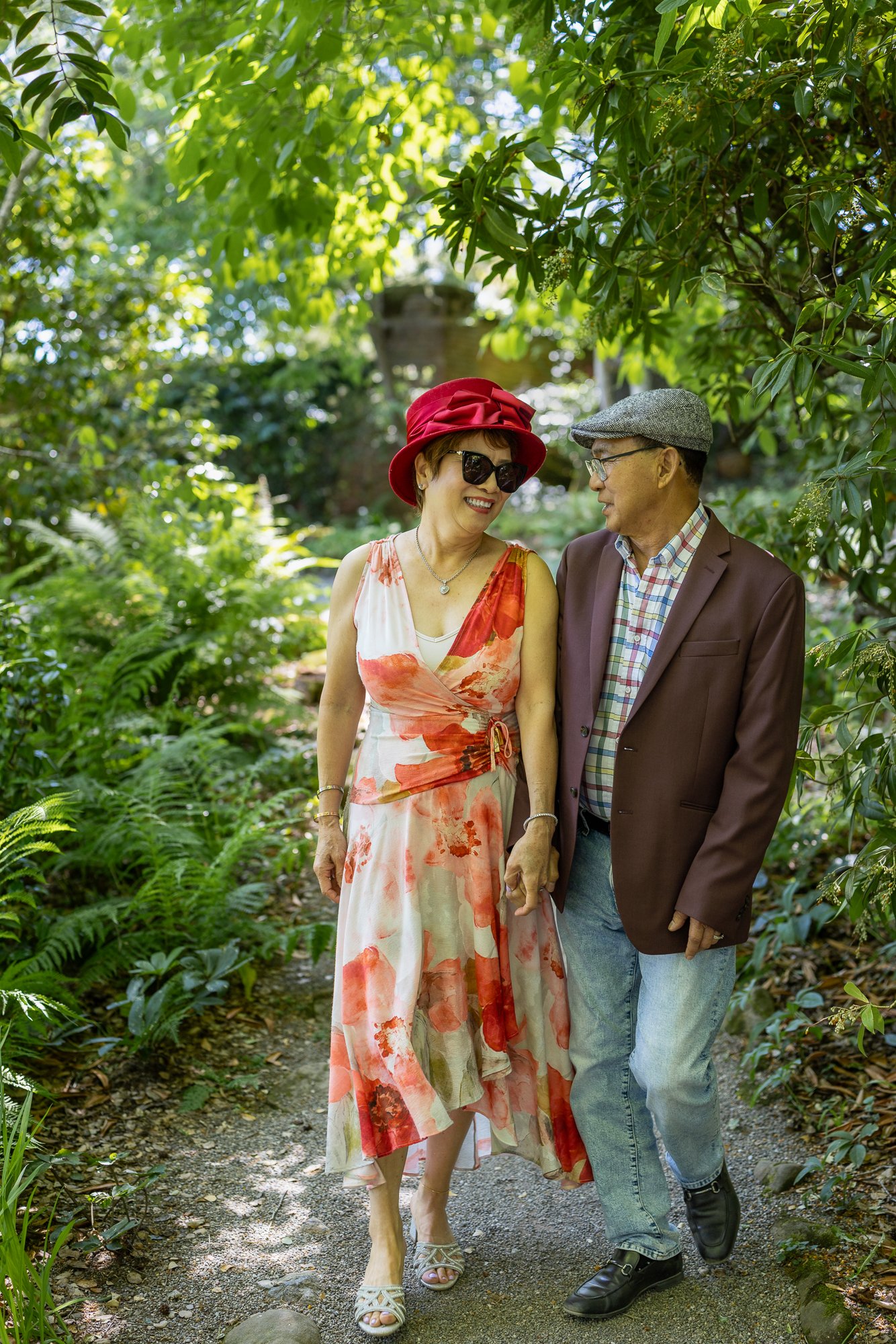 A smiling woman and a man walking together on a garden path surrounded by lush green foliage, holding hands and enjoying each other's company during the daytime.