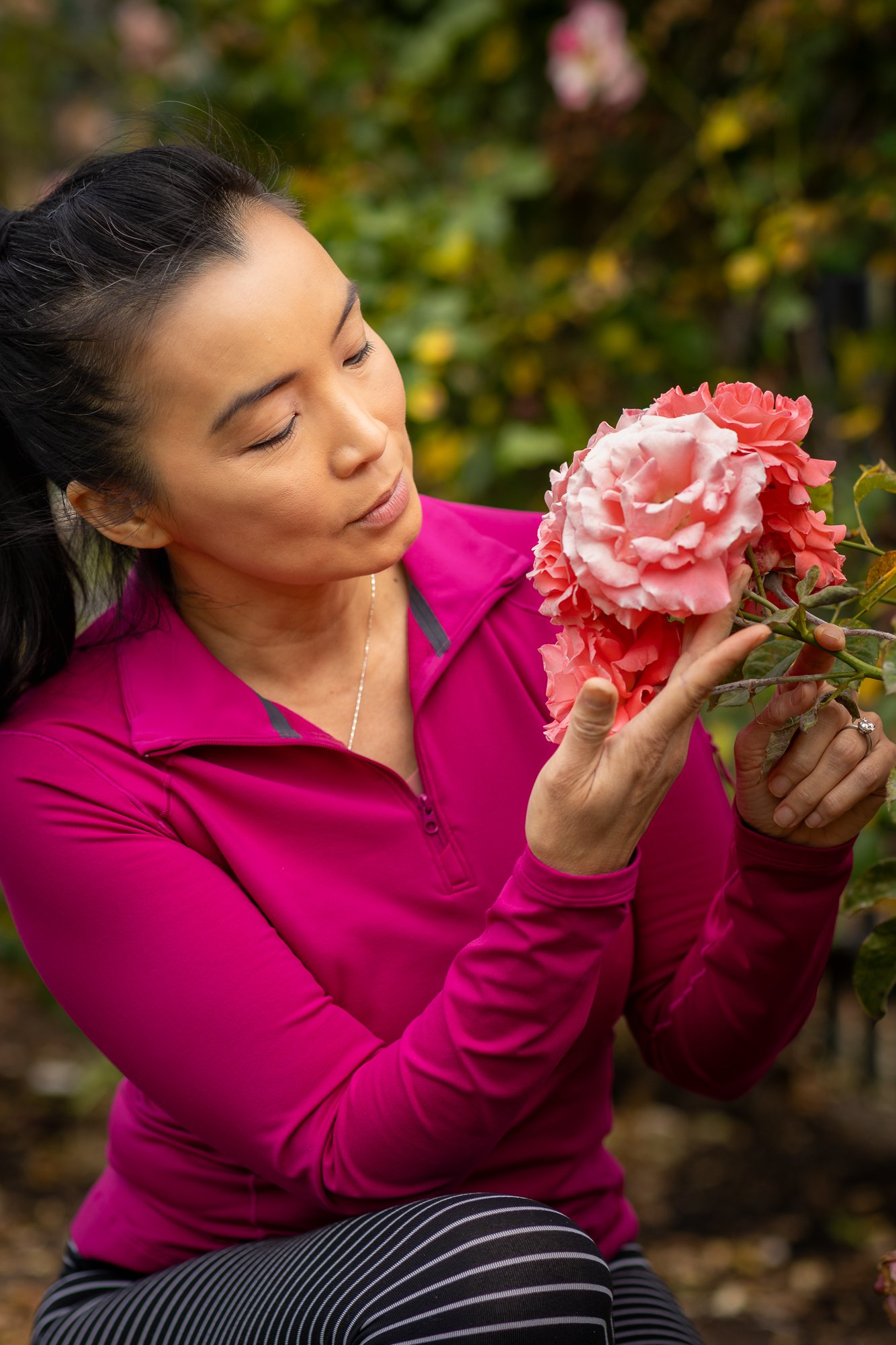 A woman with dark hair in a ponytail wearing a pink athletic jacket and black and white striped leggings is sitting outdoors, holding and inspecting pink flowers with green foliage in the background.