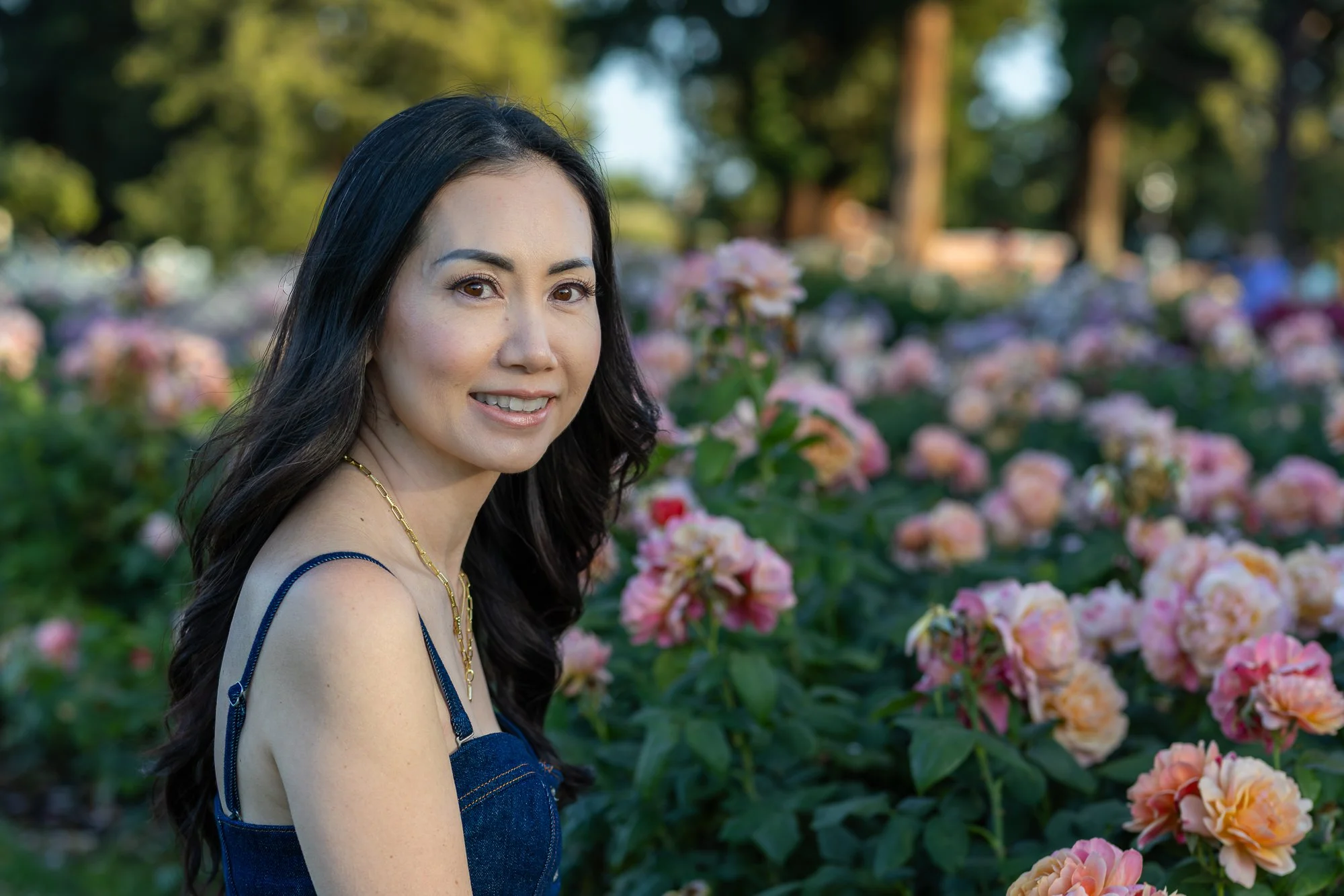 A woman with long black hair and brown eyes smiling while standing outdoors among pink and peach flowers and green trees in the background.