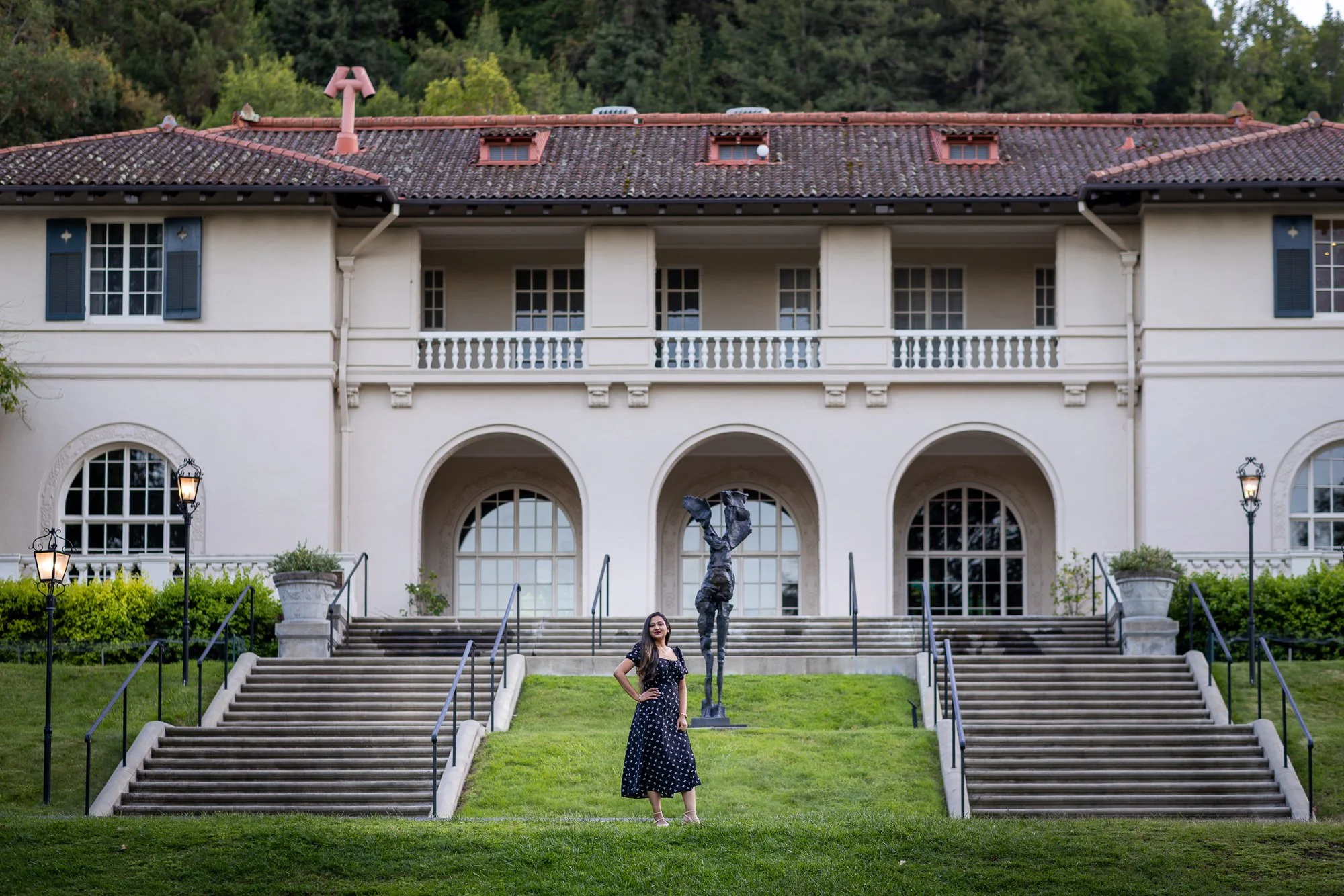 A woman in a black polka dot dress standing on grass near stairs leading up to a large white building with arched windows, columns, and blue shutters. There are street lamps and a statue in front of the building and lush green trees in the background.