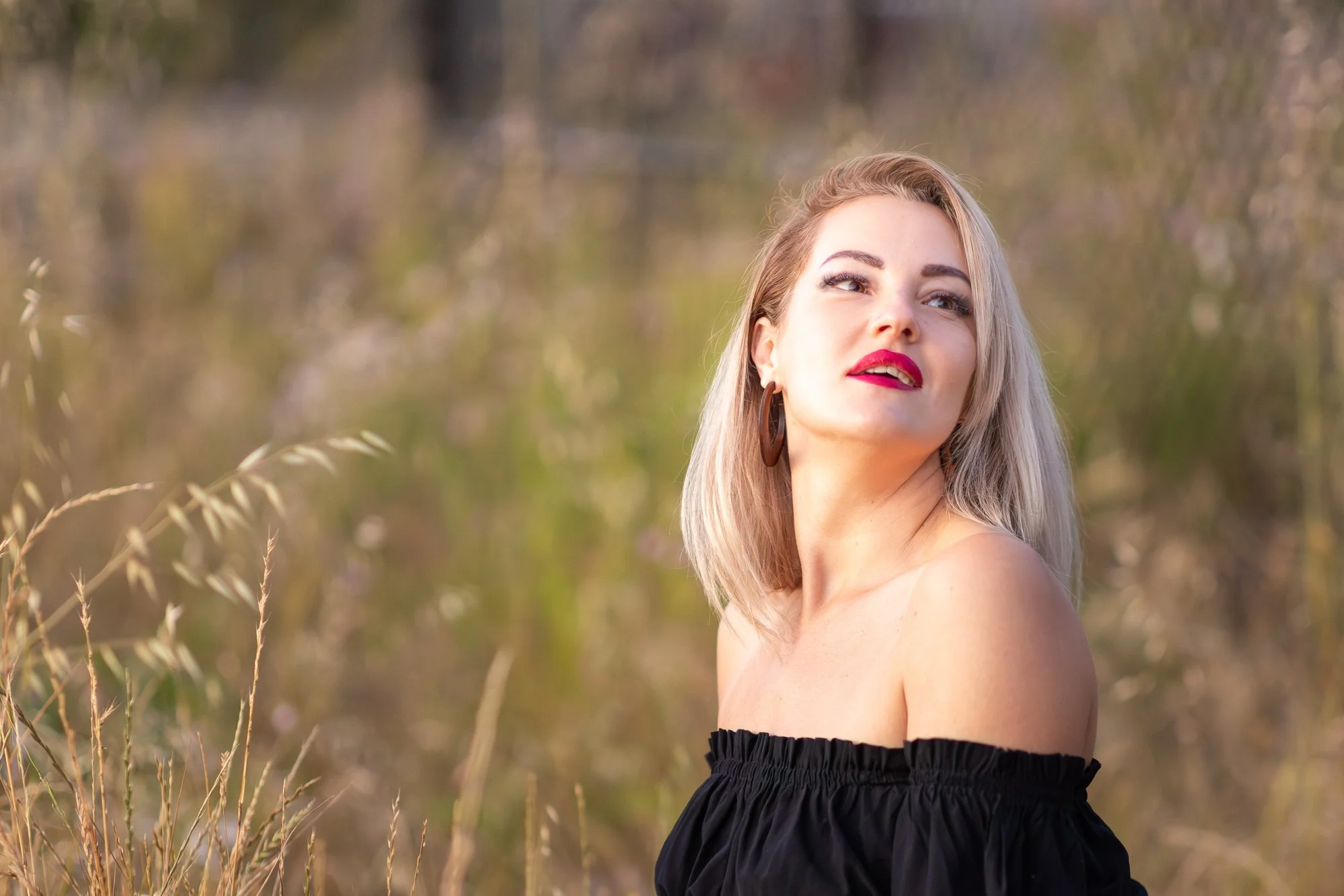 A young woman with blonde hair, wearing bold red lipstick, looking towards her left outdoors in a natural setting during daytime, with tall grass and blurred trees in the background.