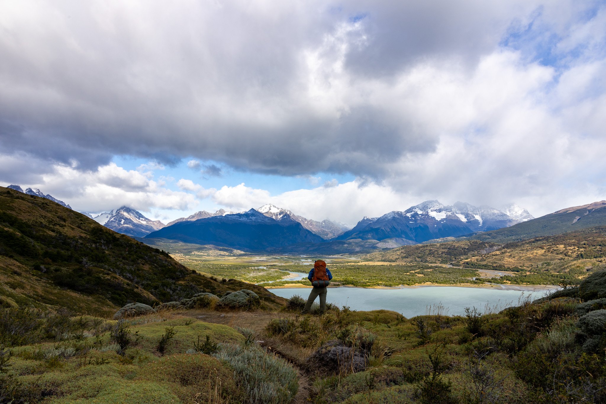 Hiker on trail above wide Patagonian valley with winding turquoise river and snow-capped mountains, Torres del Paine northern circuit