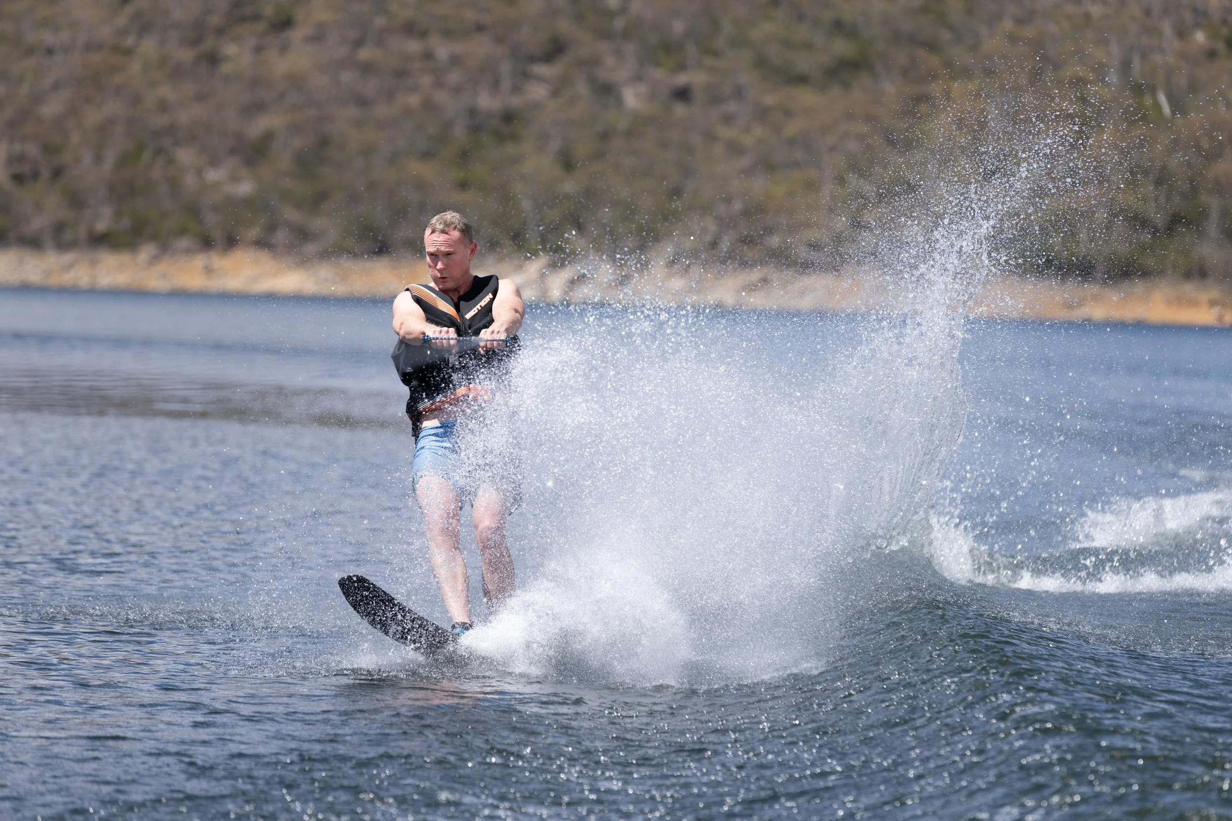 A man wake surfing on a lake, wearing a life jacket, water splashing around him as he rides on the wake.