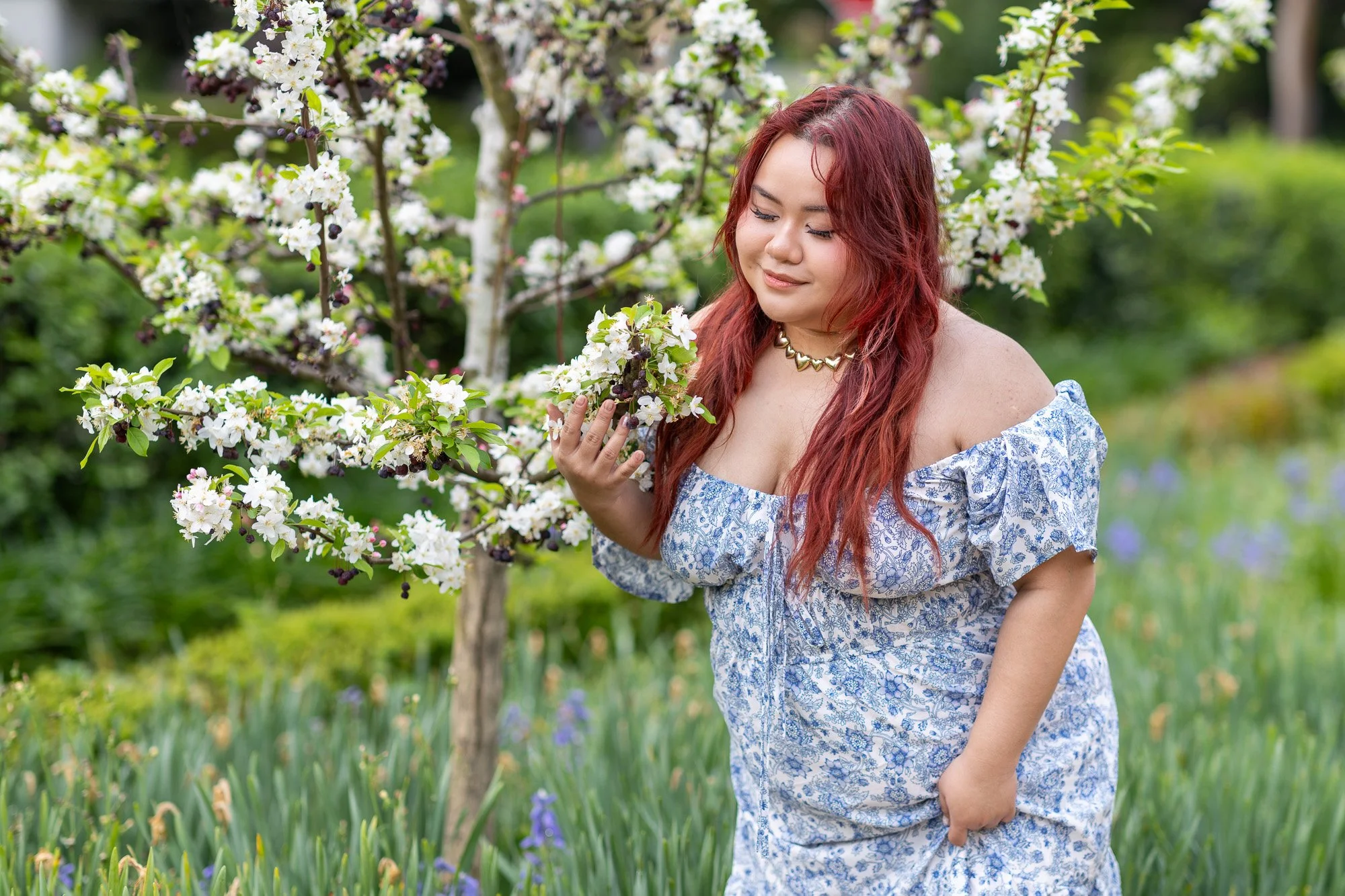 Woman in blue floral off-shoulder dress bending toward white apple blossom branch with soft downward gaze, spring orchard portrait Gamble Garden Palo Alto