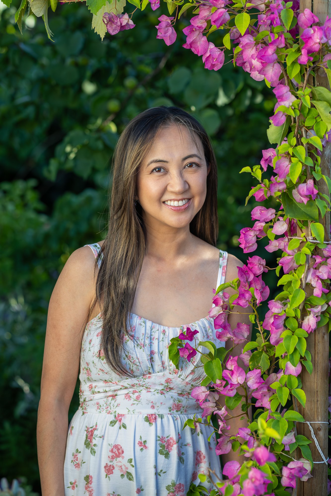 A woman with long brown hair smiling outdoors next to pink bougainvillea flowers climbing on a wooden post, with a green leafy background.