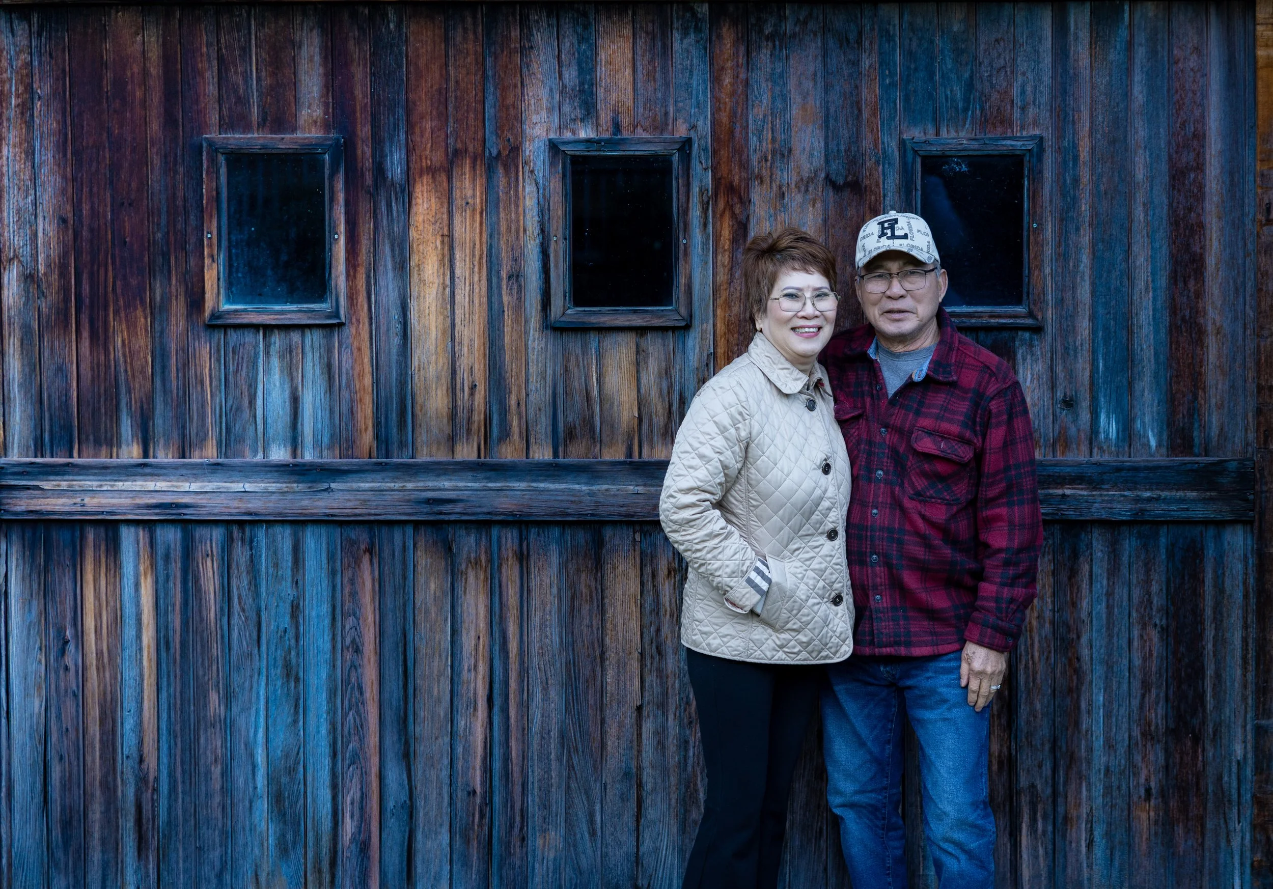 A smiling elderly woman and man standing outdoors in front of a weathered wooden wall with three small windows, the woman wearing glasses, a beige quilted jacket, and the man wearing glasses, a baseball cap, and a red plaid shirt.