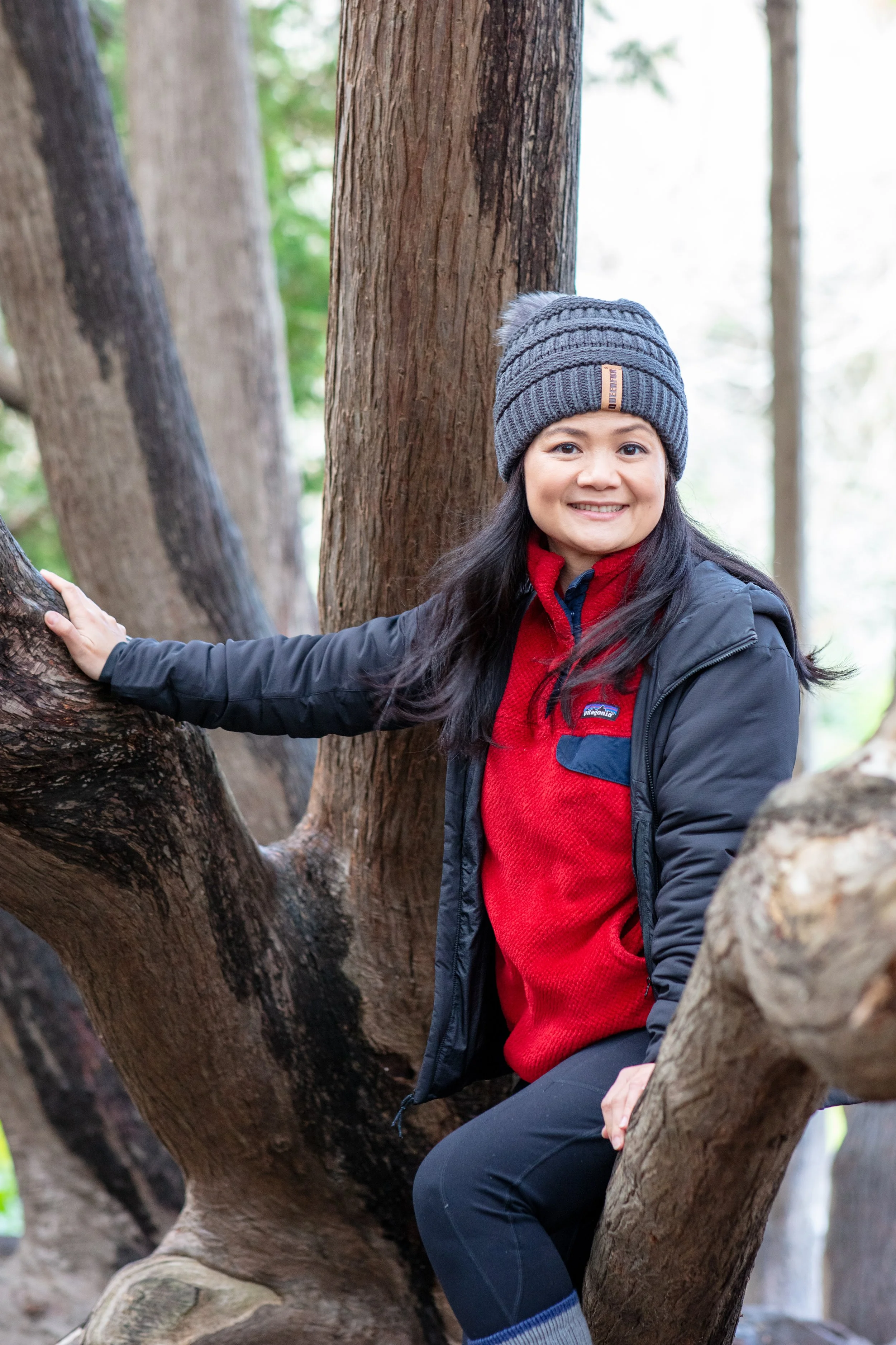 A woman wearing a gray knit hat, red fleece, black jacket, and black pants, sitting on a tree branch in a forested area, smiling at the camera.