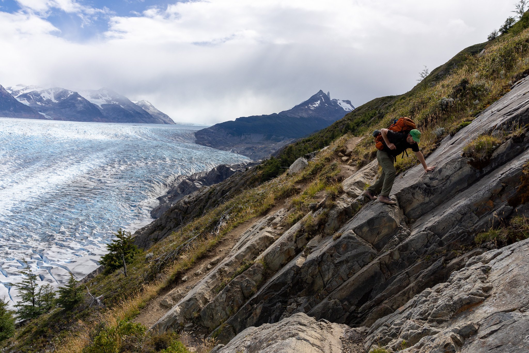 Hiker scrambling up steep exposed rock face on trail above Glacier Grey, vast blue ice field filling the frame, Torres del Paine O Circuit