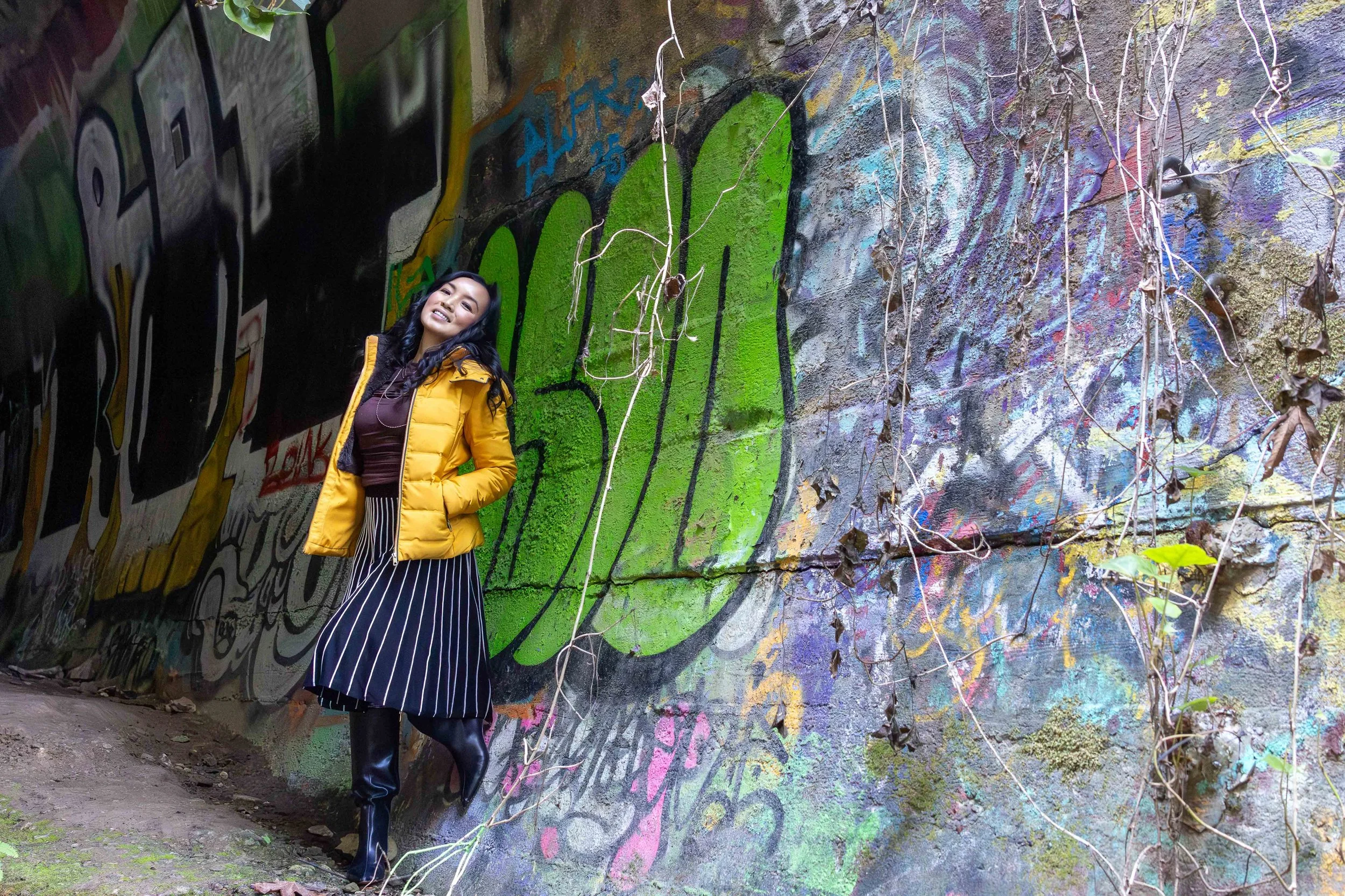 A woman standing in front of a graffitied wall, smiling, wearing a yellow puffer jacket, a dark dress with white stripes, and black knee-high boots.
