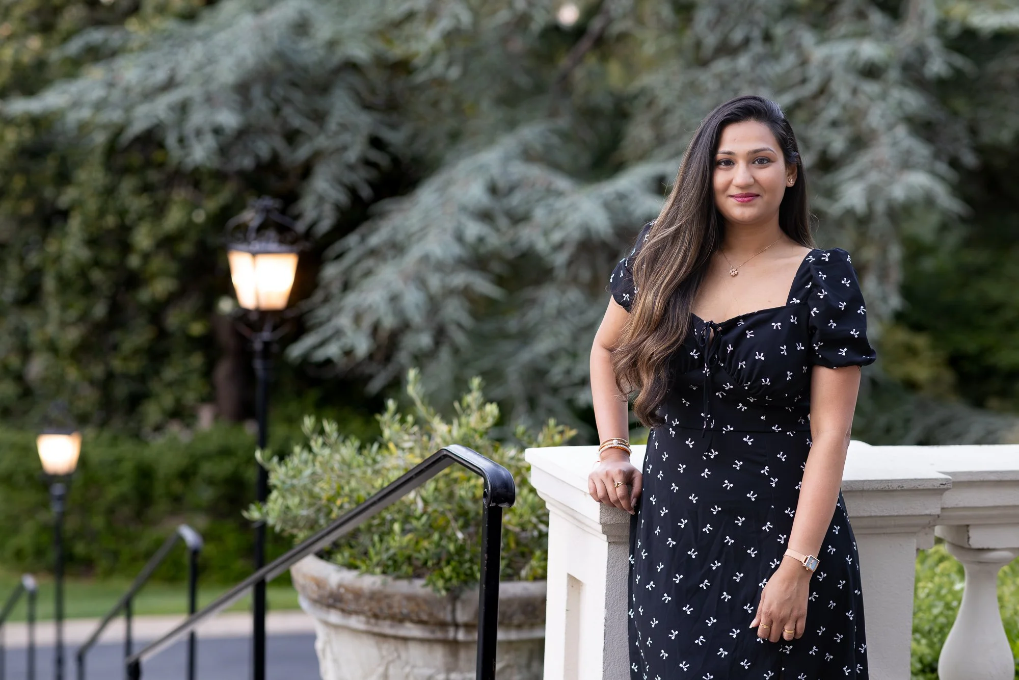 A woman with long dark hair and a black dress with white bows, standing outdoors near a white railing and a large flower pot, with trees and garden lights in the background.