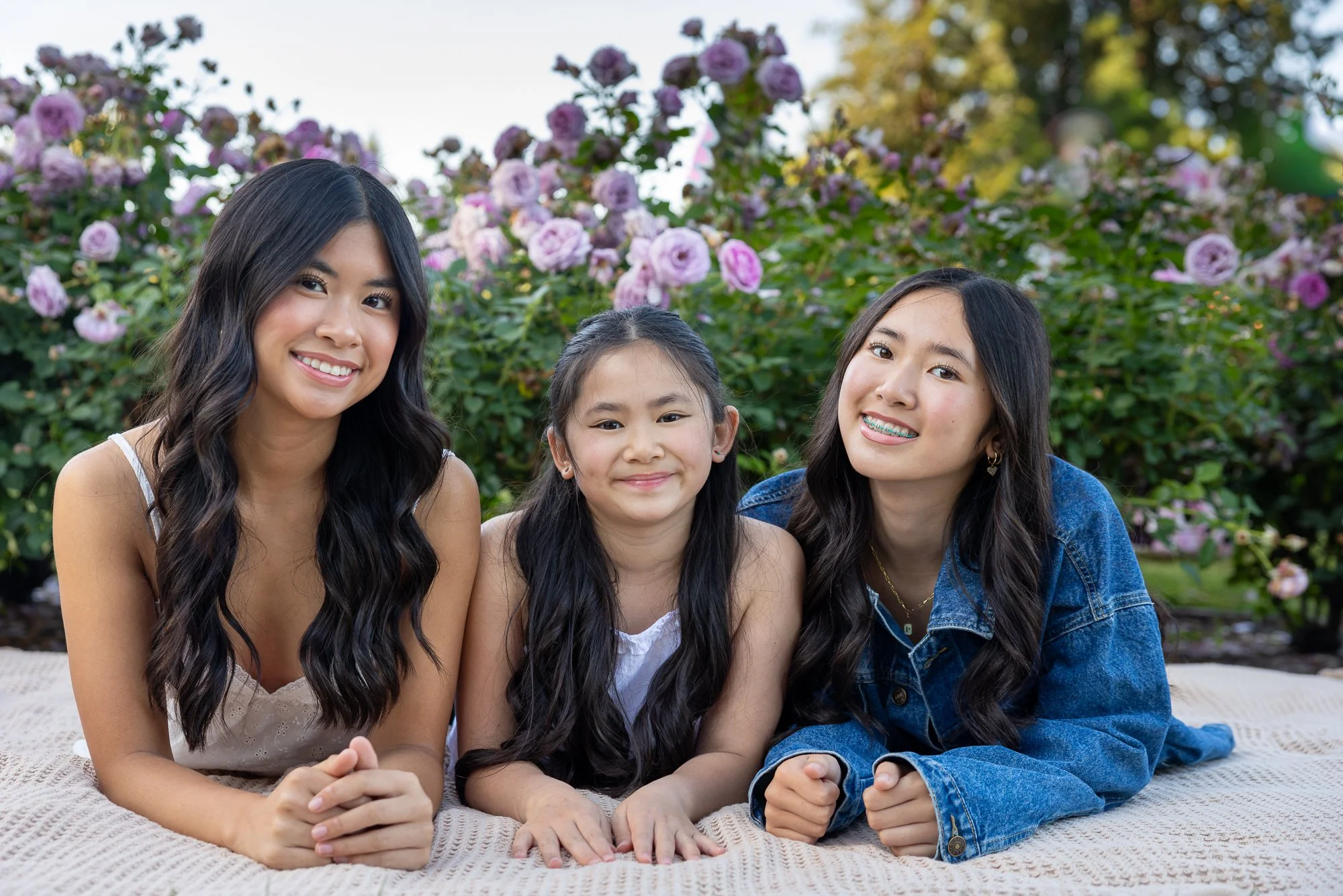 Three girls sitting on a blanket outdoors with pink roses in the background.