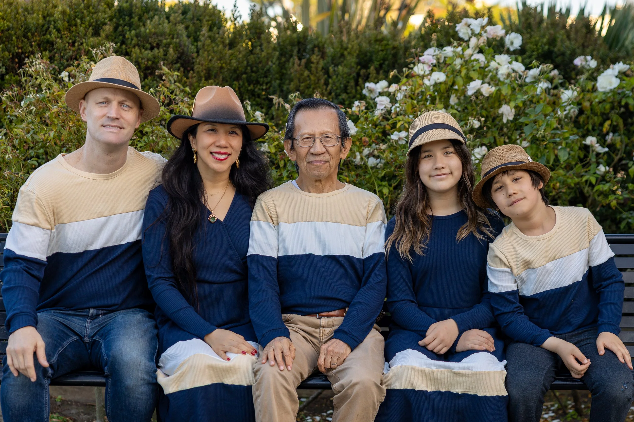 A multigenerational family sitting on a park bench in front of flowering bushes, smiling at the camera.