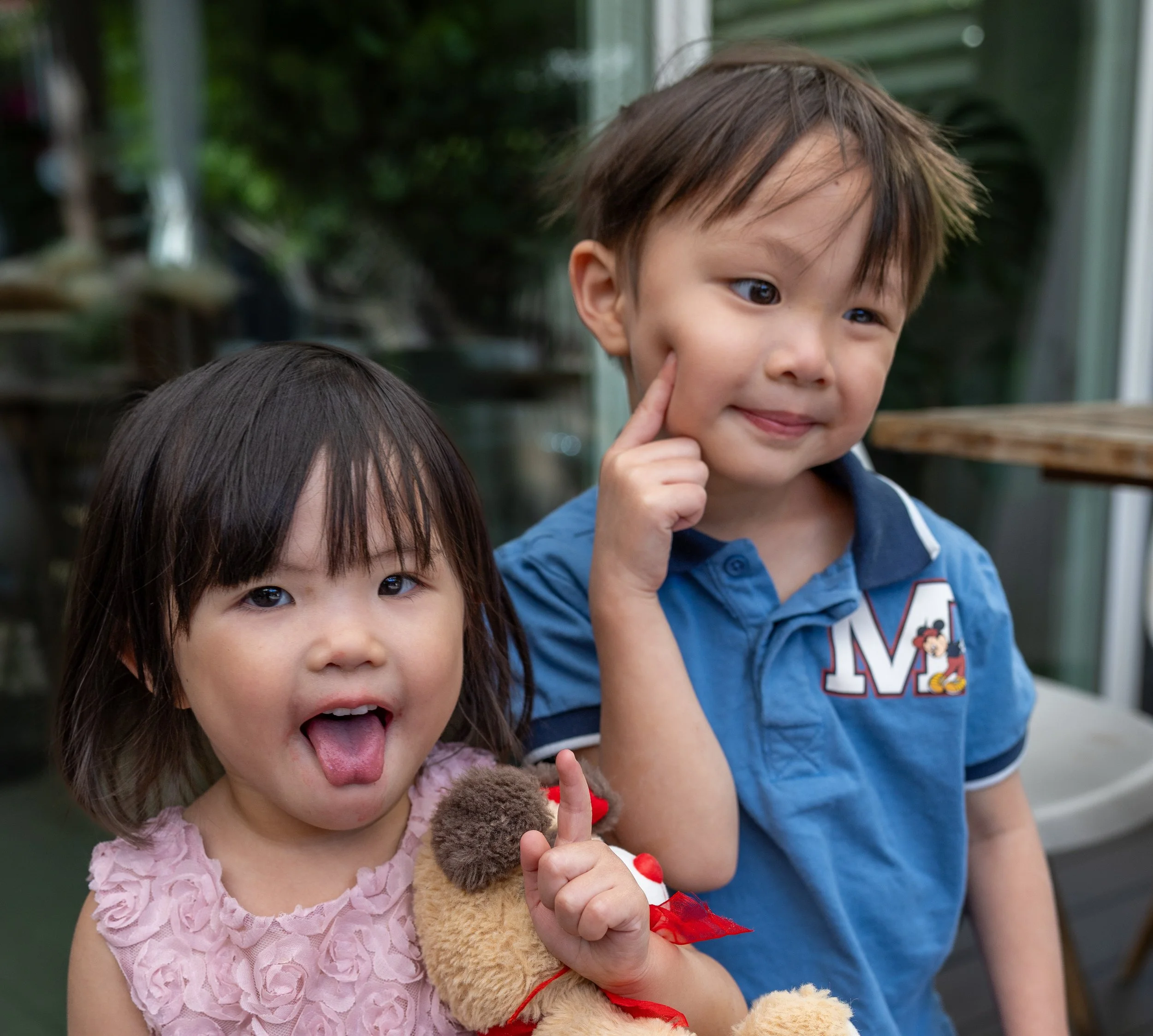 Two young children, a girl with dark hair and a boy with light brown hair, posing outdoors. The girl is sticking out her tongue and holding a teddy bear, while the boy is smiling and pointing to his cheek.