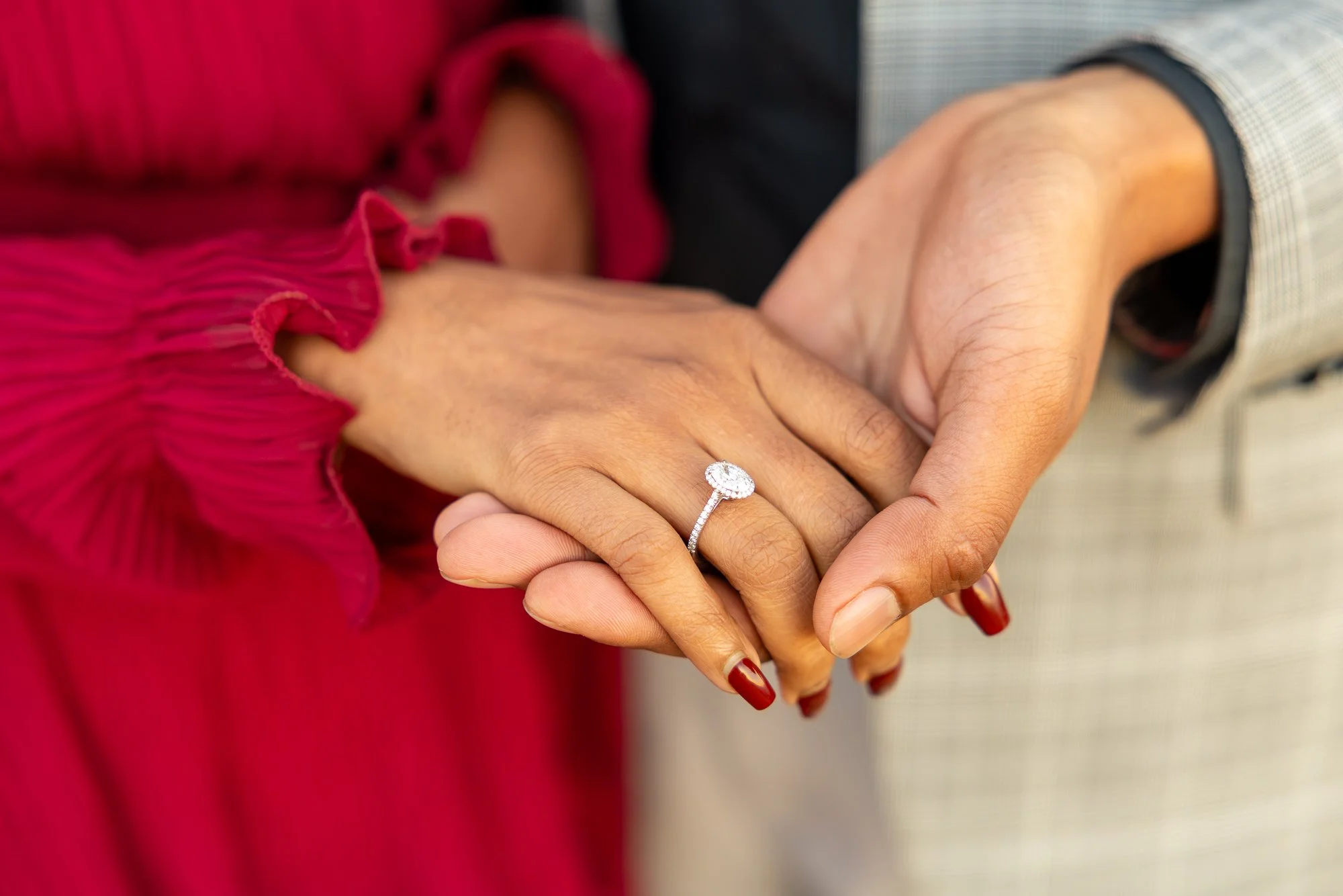 Close-up of a couple holding hands, with the woman wearing a red outfit with ruffled cuffs and displaying an engagement ring. The man is wearing a checkered shirt, and the focus is on their hands and the ring.