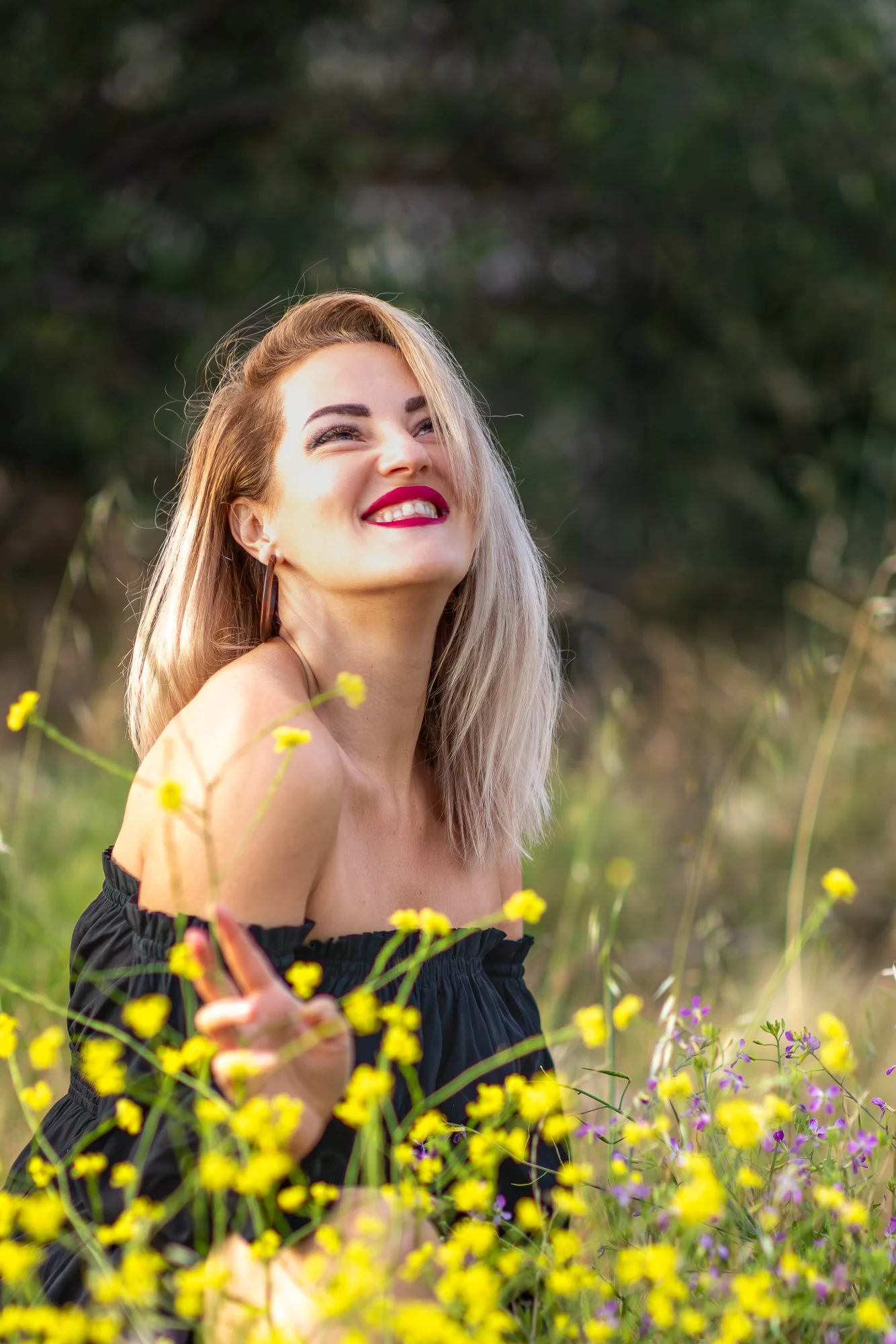 Smiling woman with blonde hair wearing a black off-shoulder dress, standing among yellow and purple wildflowers in a field.