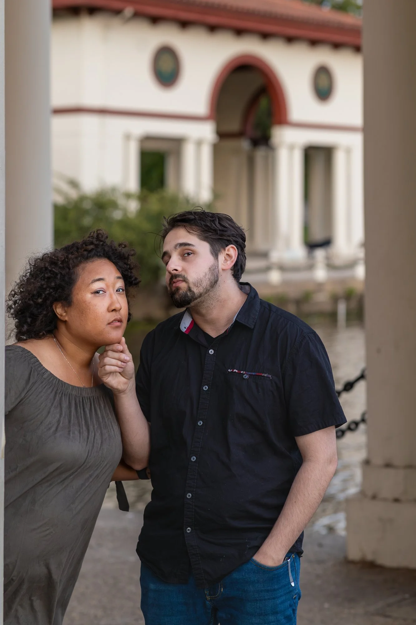 Two people posing outdoors near a waterfront with a building in the background, one with curly hair and the other with straight hair, both with serious expressions.