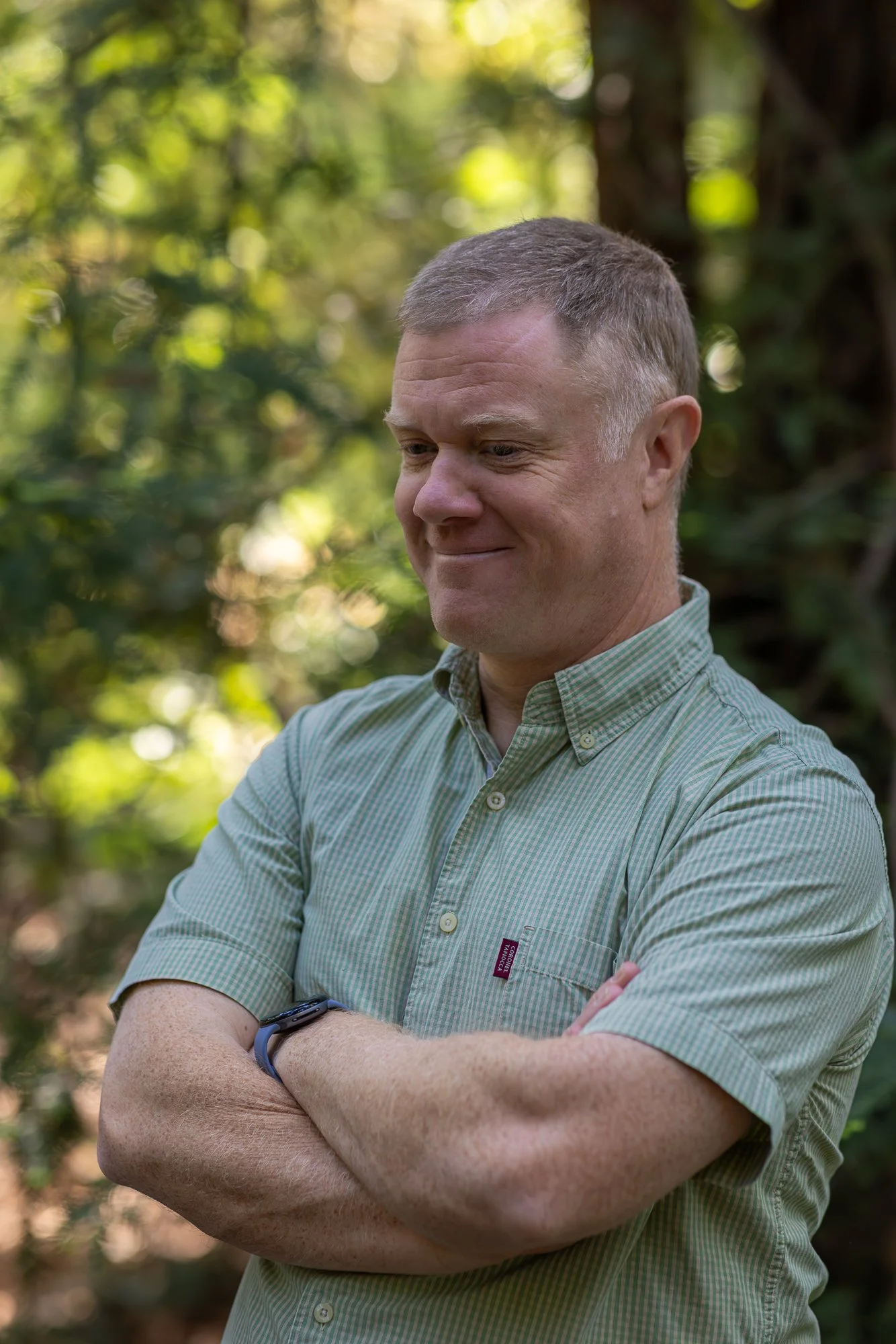 A middle-aged man with short gray hair, wearing a green checkered shirt, standing outdoors with arms crossed, smiling slightly against a background of blurred green foliage.