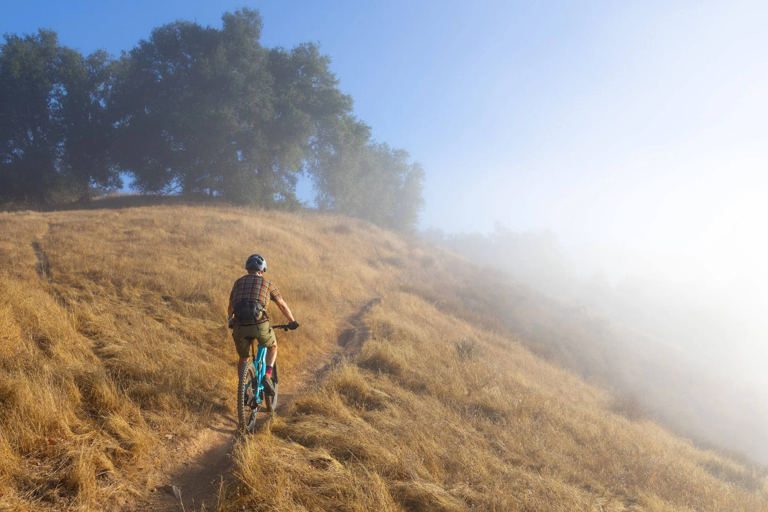 A person mountain biking on a narrow dirt trail on a grassy hillside, with fog on the right and some trees at the top of the hill under a clear blue sky.