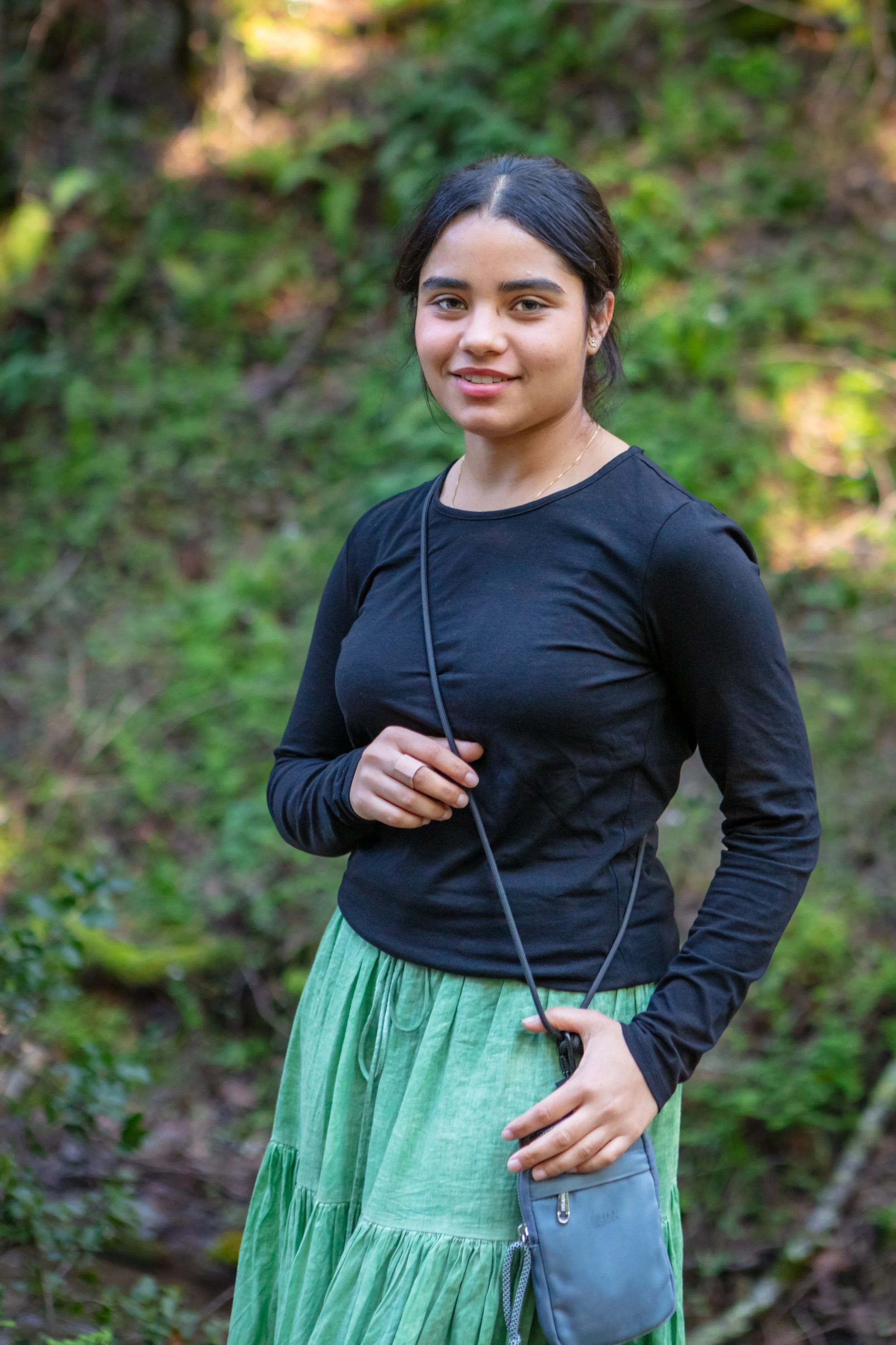 A young woman with dark hair, wearing a black long-sleeve shirt and a green skirt, standing outdoors in a forested area, holding a small bag and looking at the camera.