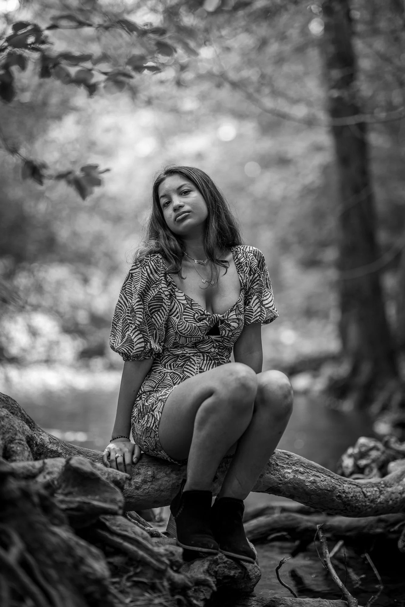 Black and white photo of a young woman sitting on a fallen tree by a river in a forested area, looking at the camera with a serious expression.