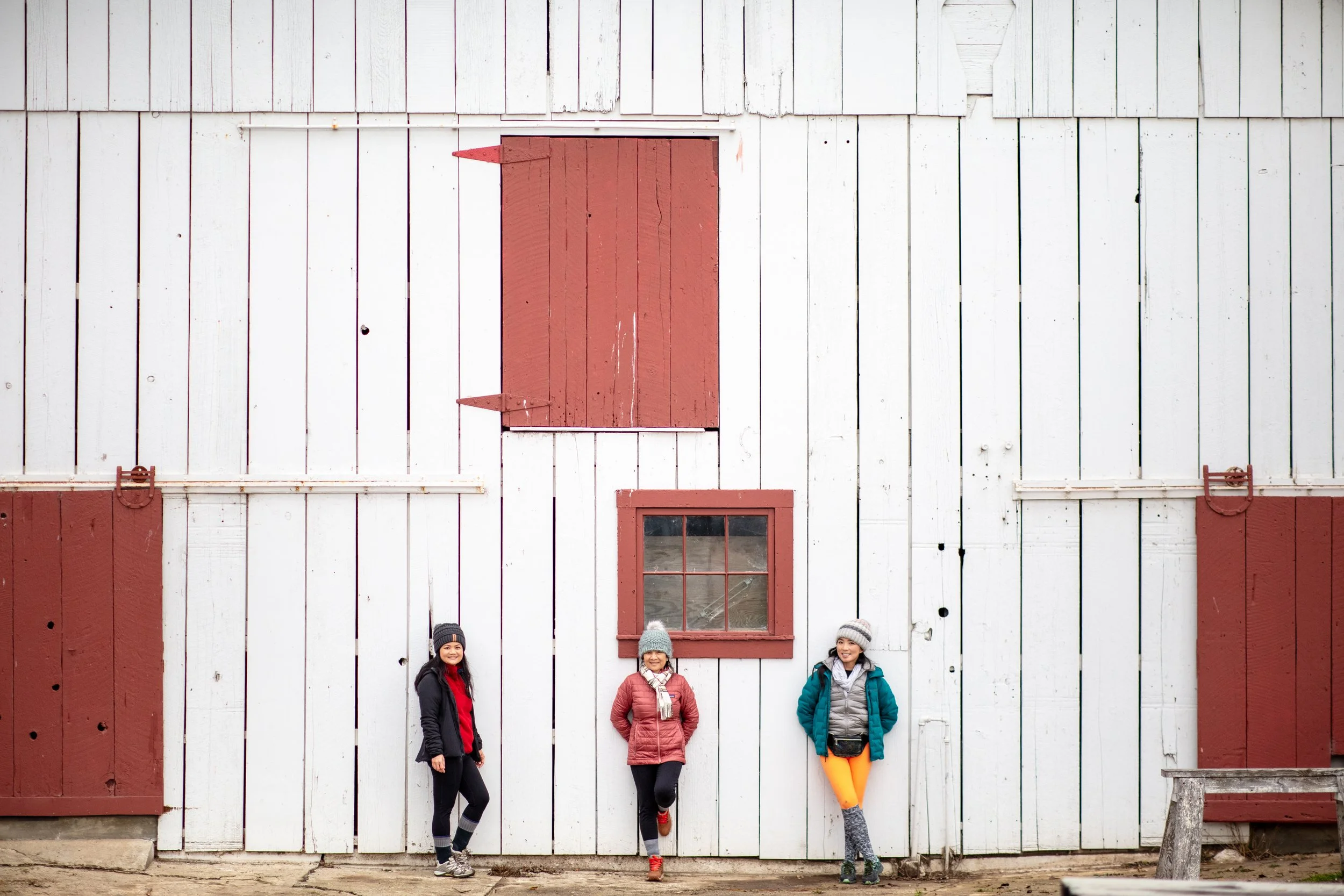 Three women standing in front of a large white barn with red accents, wearing winter jackets and hats.