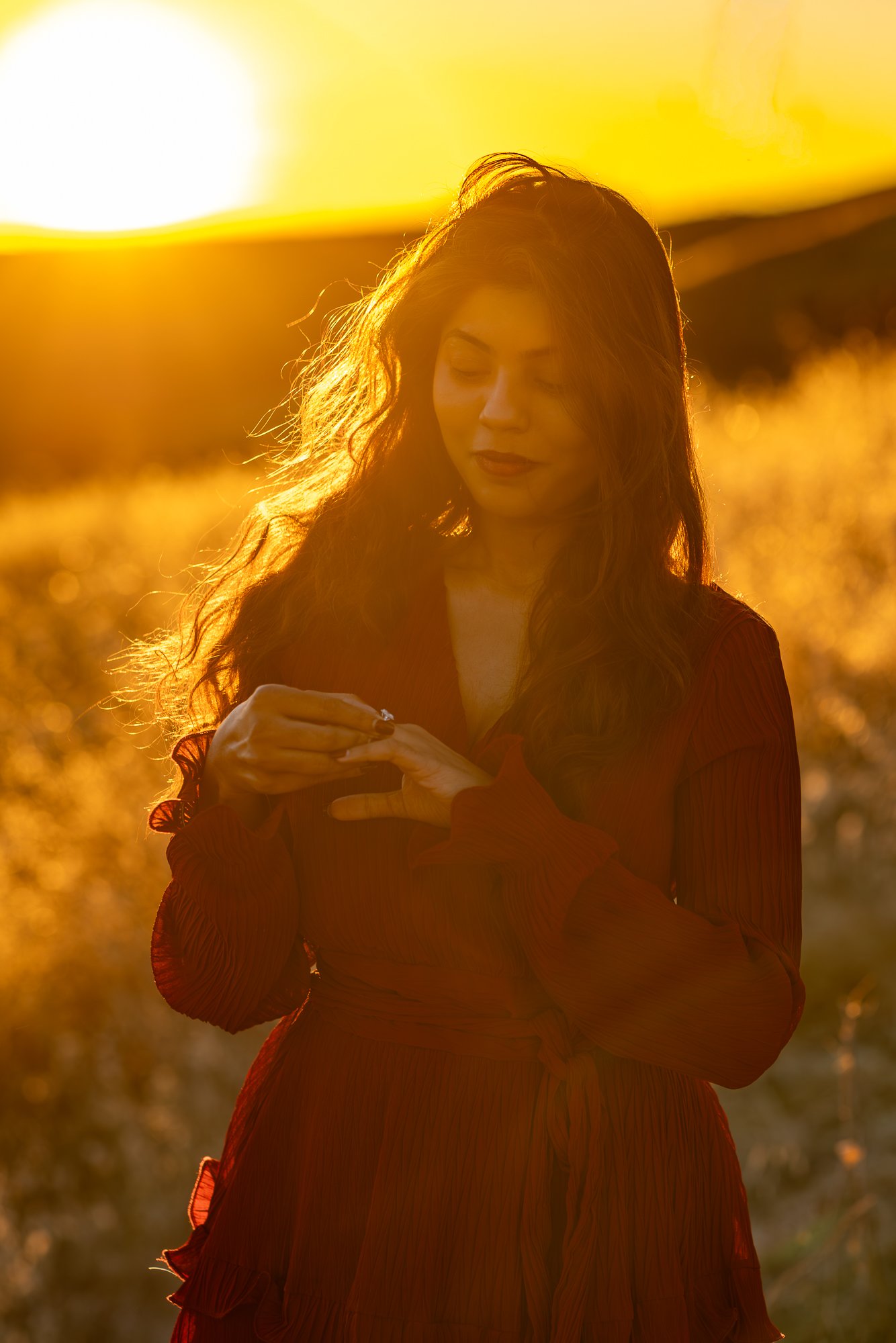 A woman with long hair standing outdoors during sunset, wearing a red dress, with sunlight behind her creating a warm glow.
