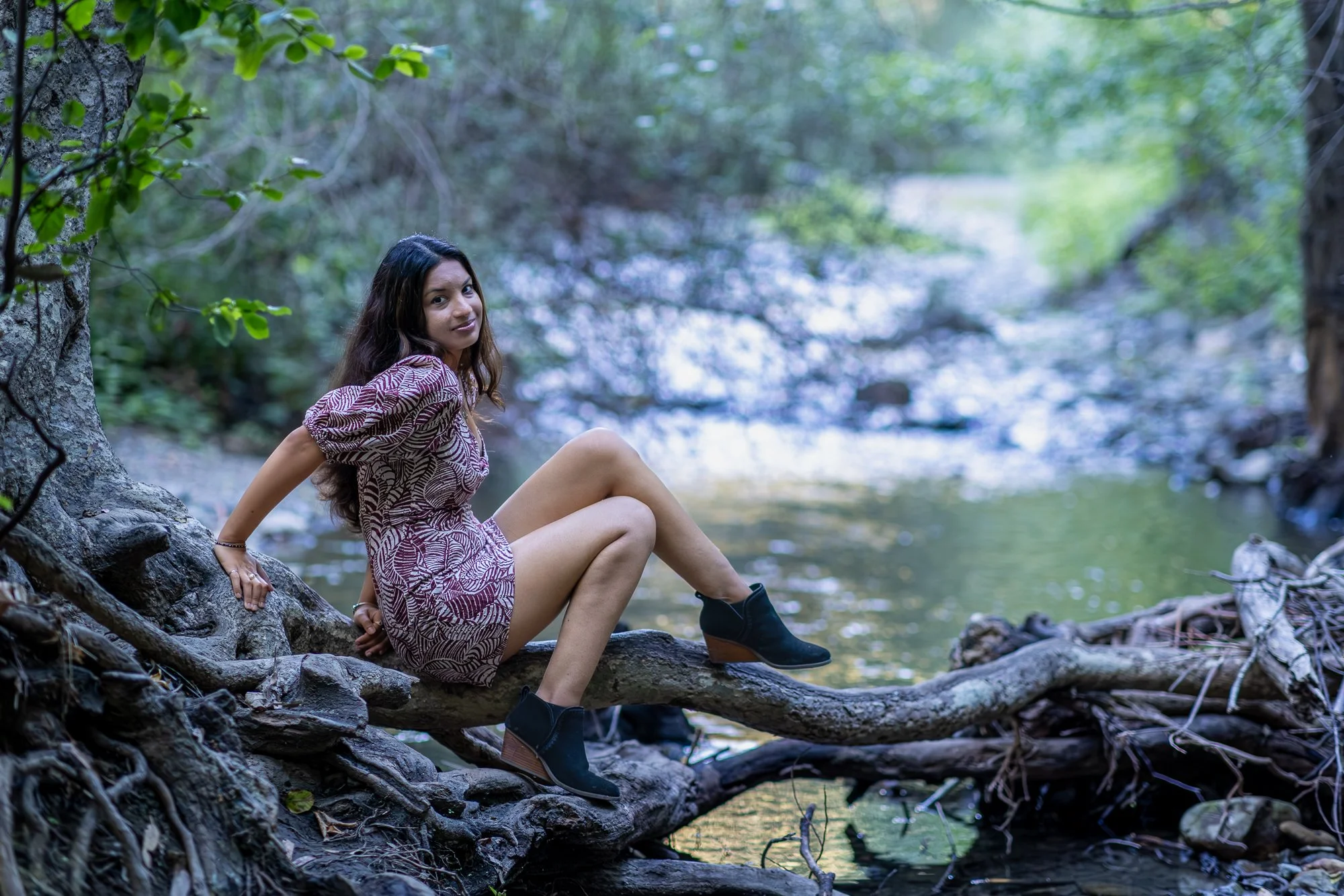 A woman sitting on a fallen tree trunk by a river in a lush green forest, looking at the camera with a slight smile.