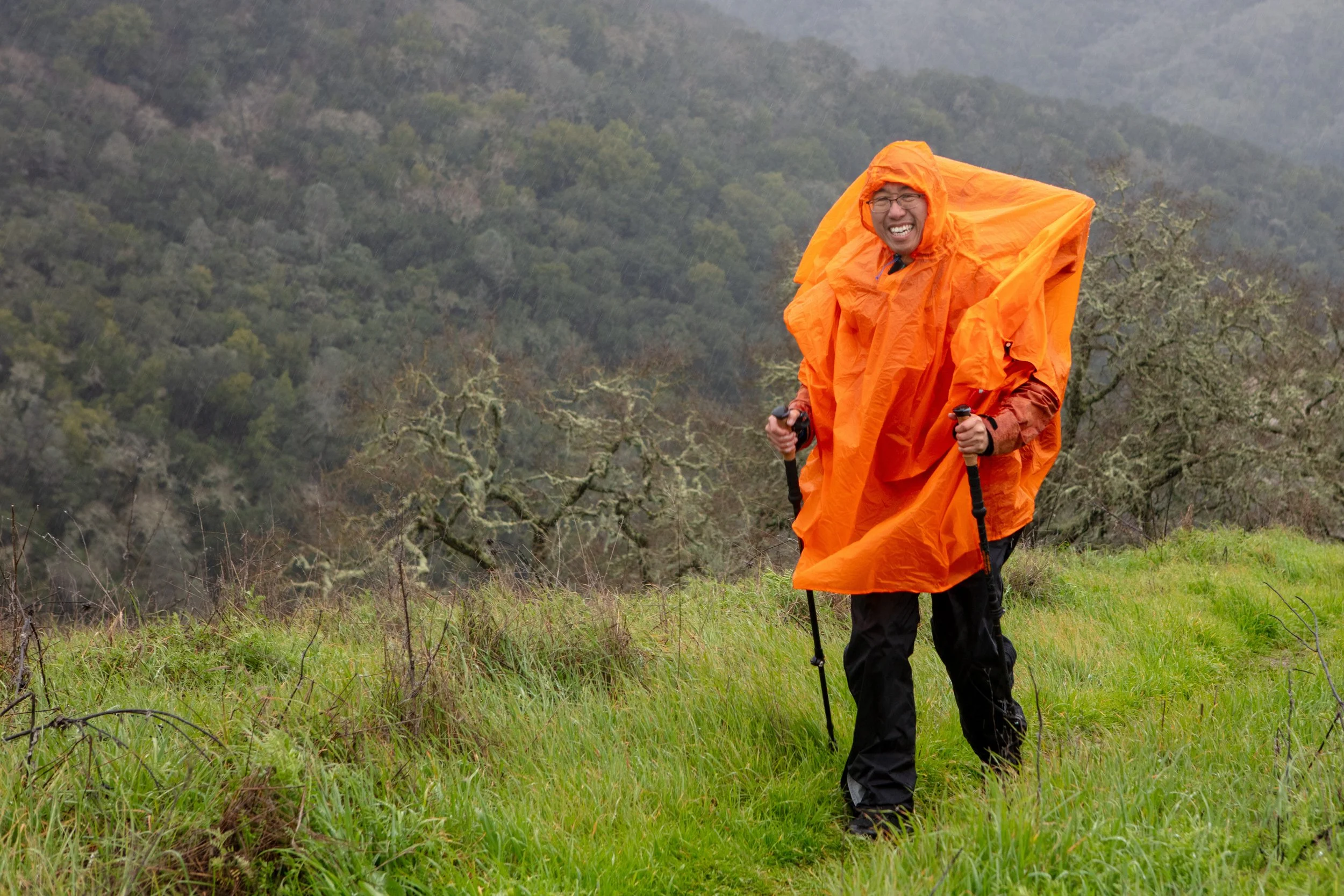 A man dressed in an orange rain poncho and black outdoor clothing hiking on a grassy trail in a misty, wooded area, smiling at the camera.