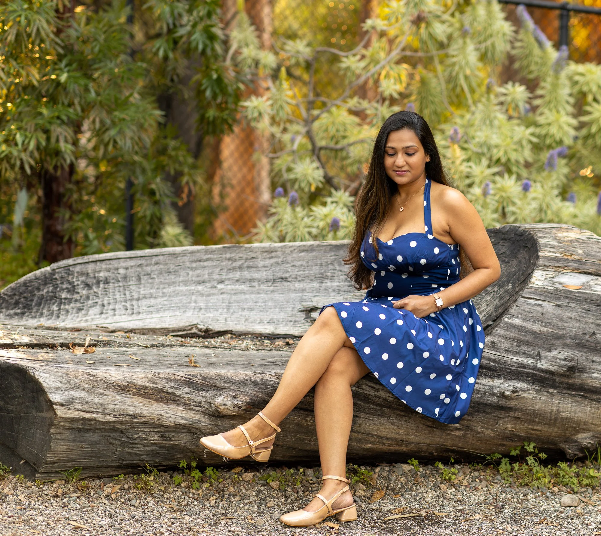 A woman in a blue polka dot dress with beige shoes sitting on a large weathered tree trunk in a natural outdoor setting with green and yellow foliage.