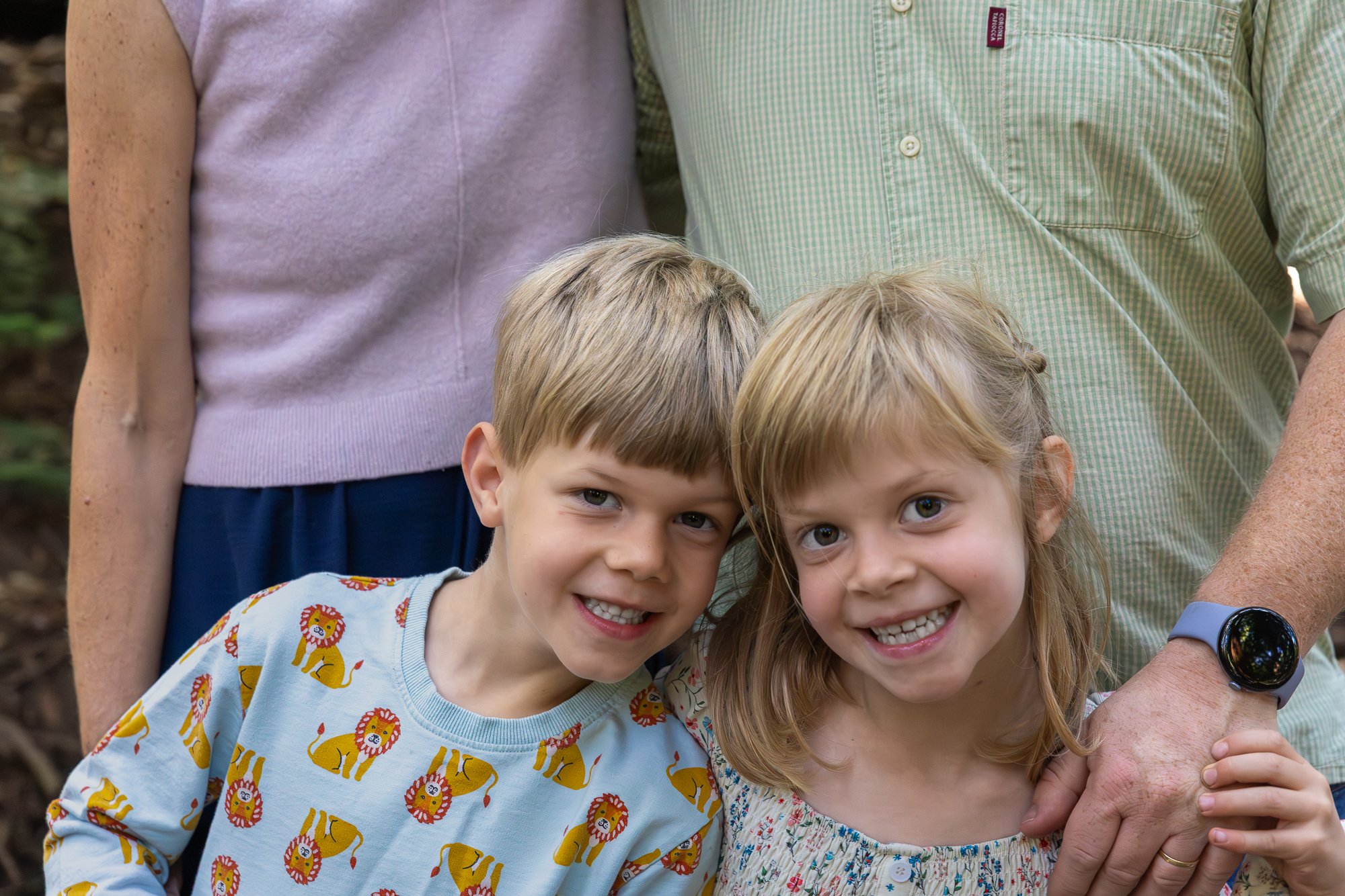 Two young children, a boy and a girl, smiling and standing close together outdoors. The boy is wearing a shirt with a lion print, and the girl is wearing a dress with a floral pattern. They are surrounded by adults, with parts of their torsos and arm