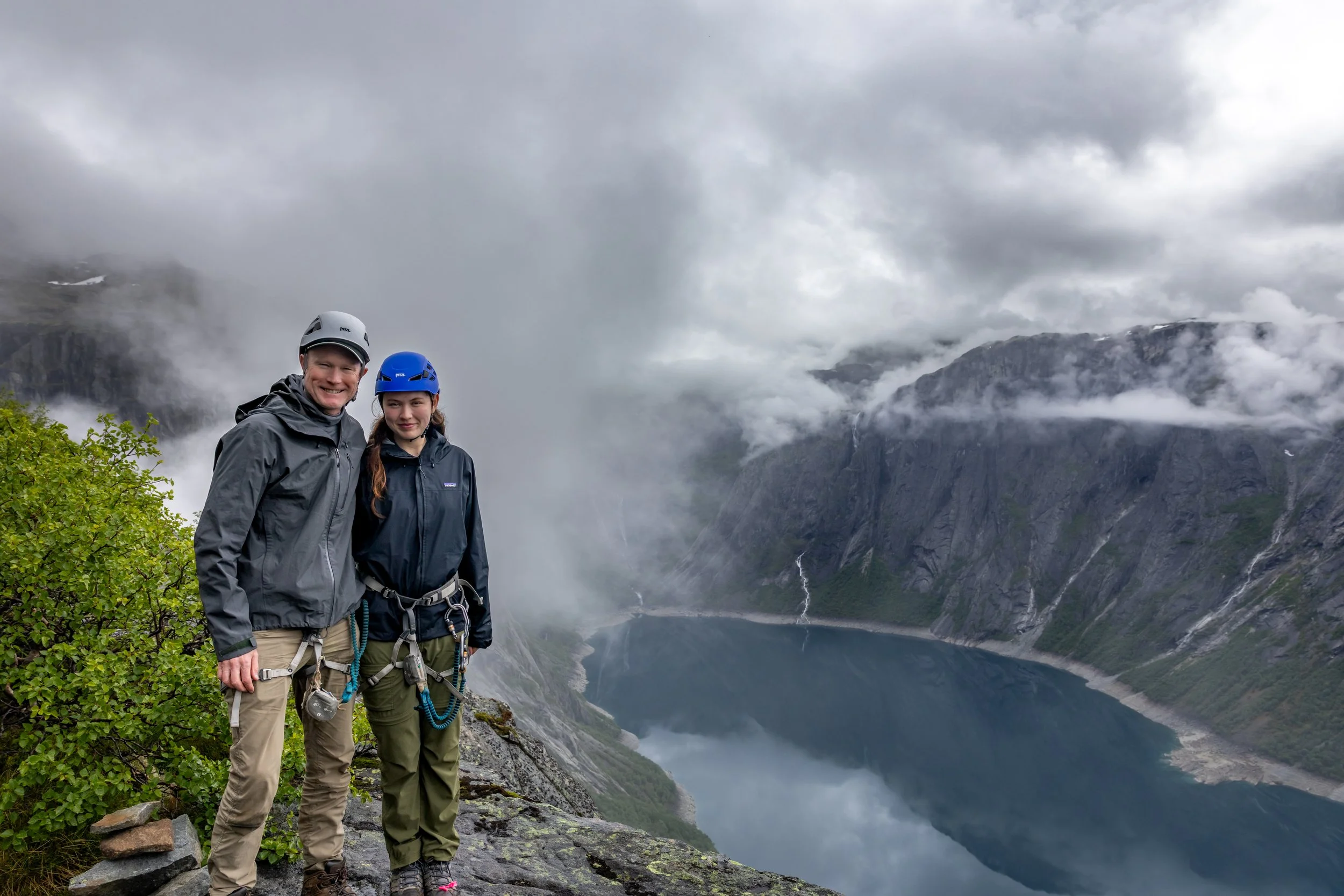 Two hikers standing together on a trail with the fjord and mountains behind them.