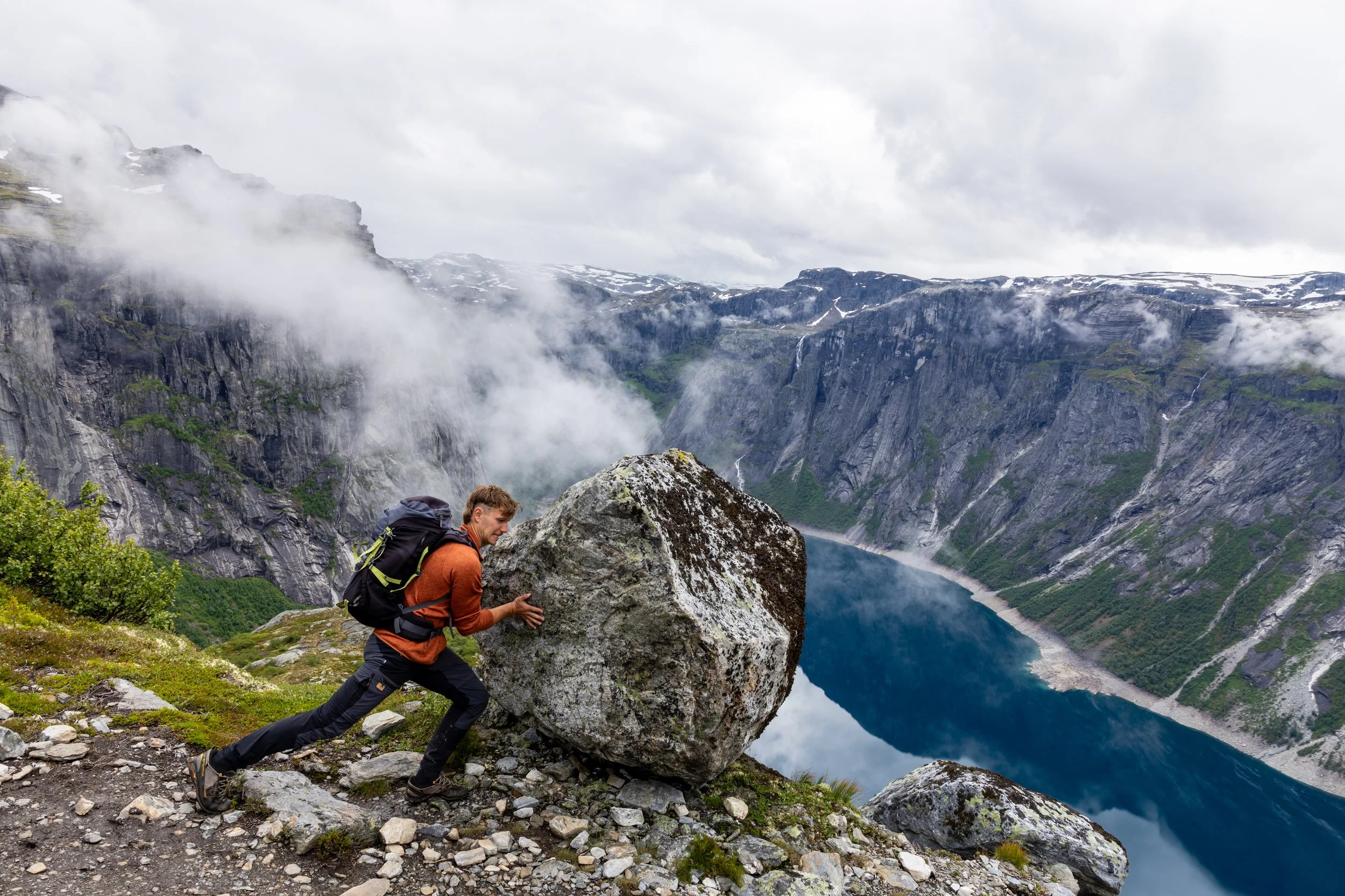 Hiker pushing a large boulder on a mountain trail overlooking a fjord with steep cliffs, clouds, and mist.