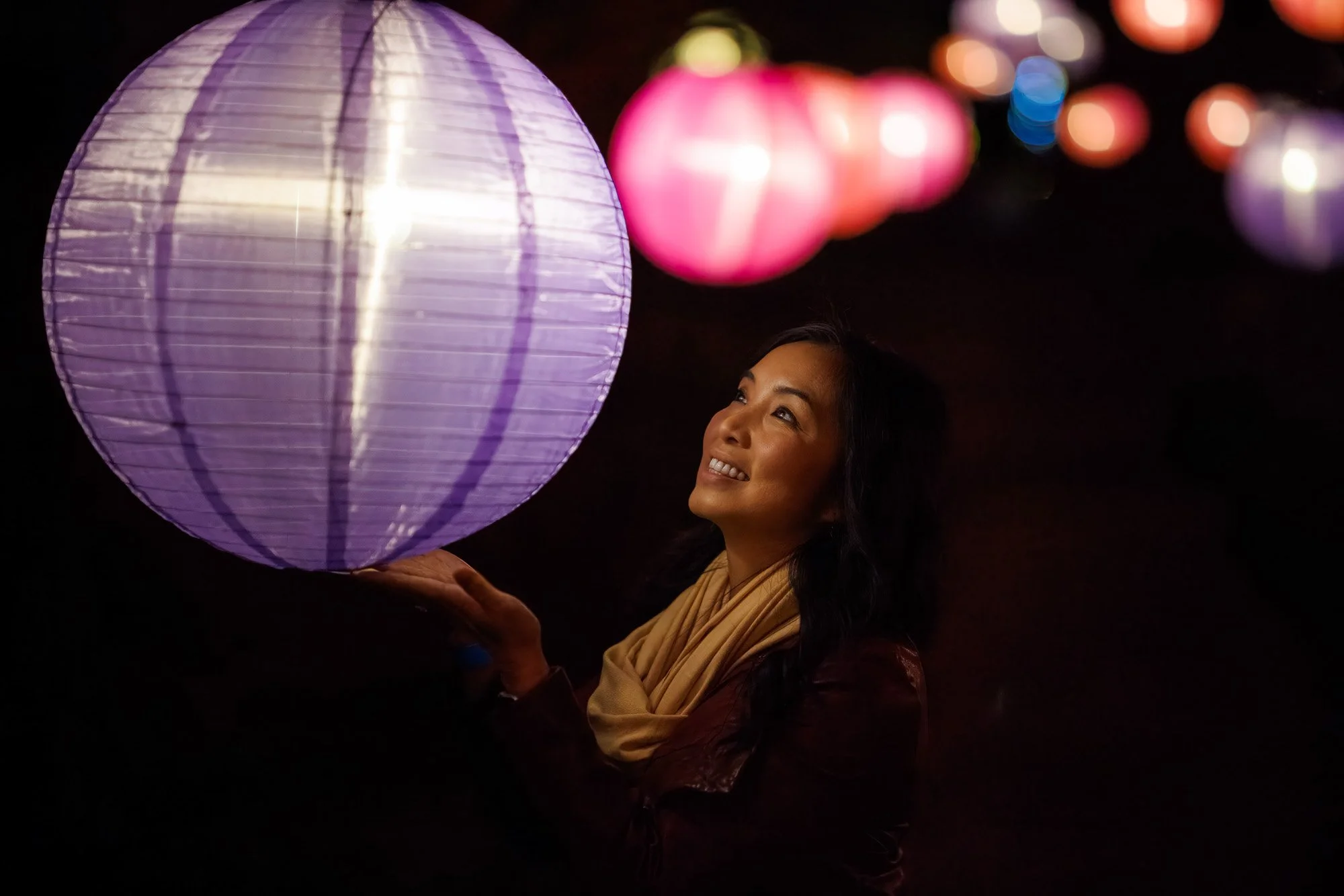 A woman smiling and looking upward while holding a large purple paper lantern at night, with colorful blurred lanterns in the background.