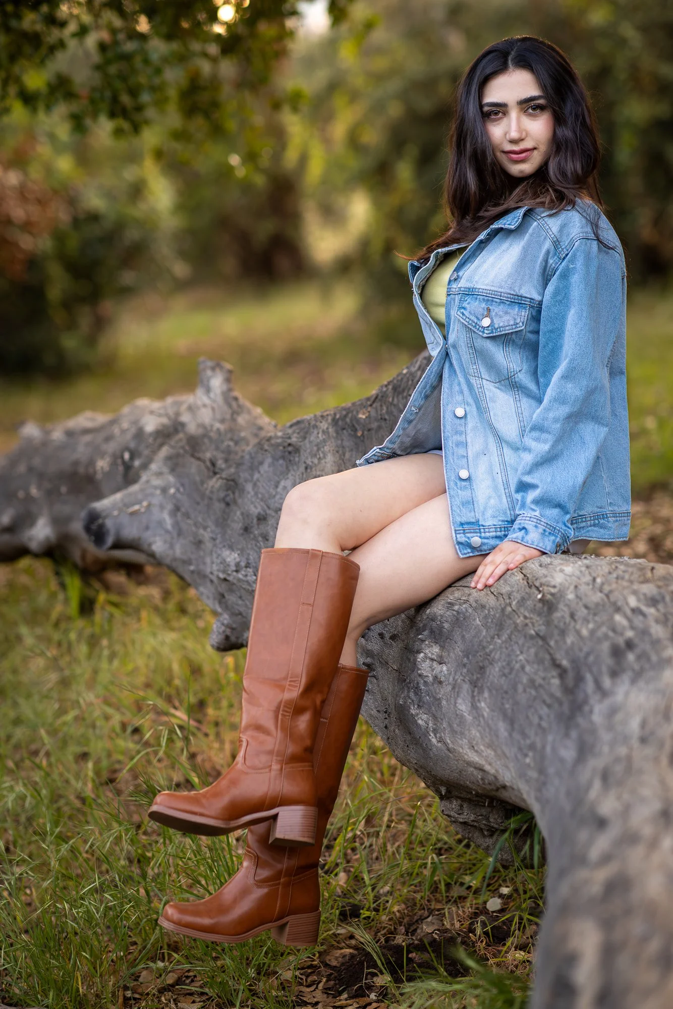 Woman in denim jacket sitting on fallen log with warm green tree canopy bokeh at Stanford Cactus Garden, natural light lifestyle portrait