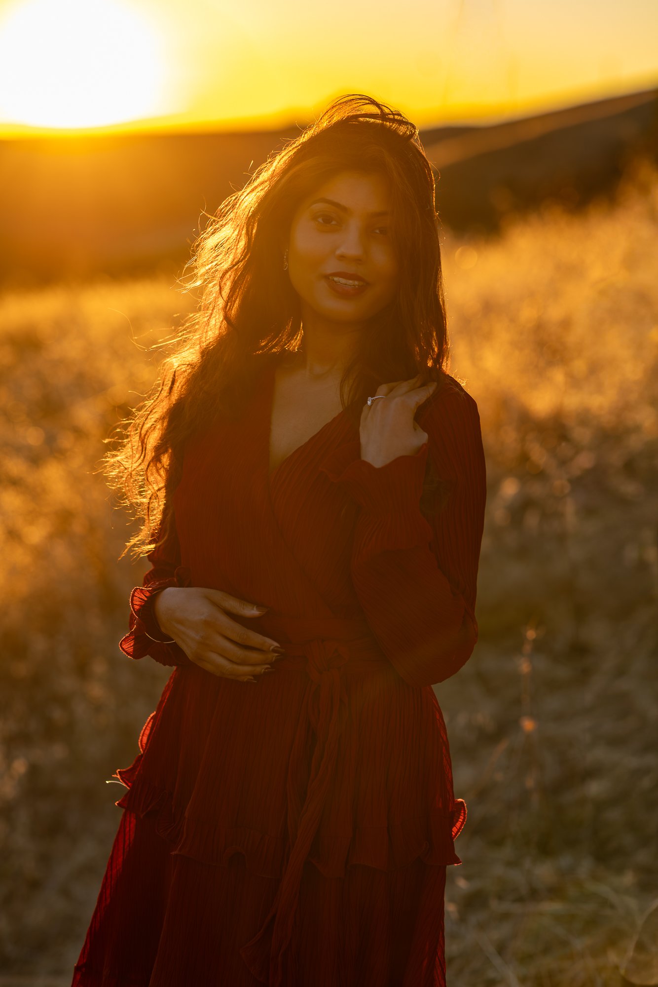 A woman with long hair standing outdoors at sunset, wearing a red dress and looking at the camera.