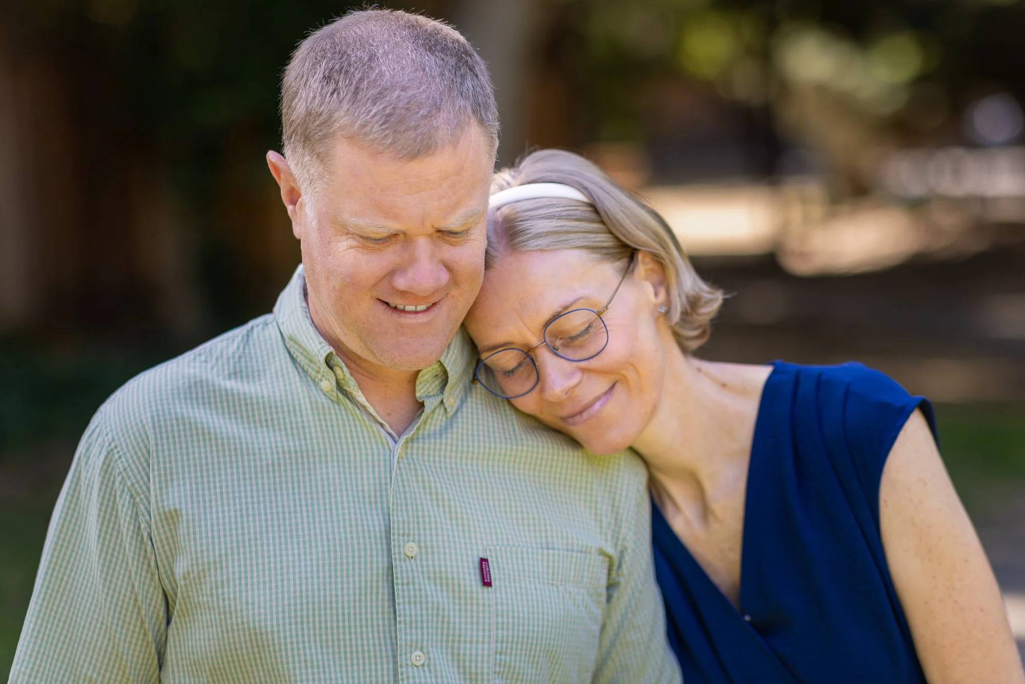 An elderly man and woman sharing a tender moment outdoors, with the woman resting her head on the man's shoulder, both appearing peaceful and affectionate.